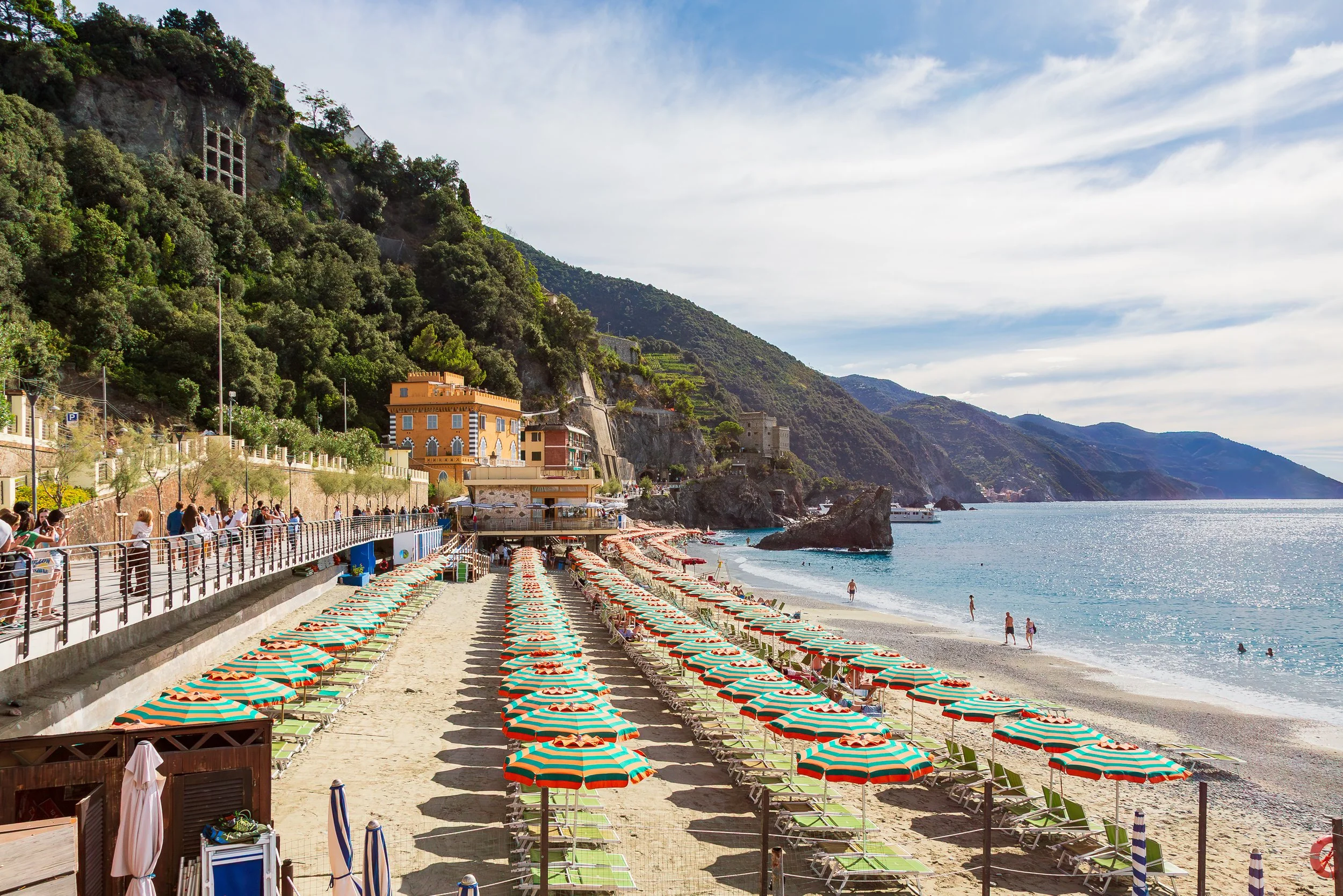 A scenic beach with colorful striped umbrellas and lounge chairs, overlooking the calm sea with rocky cliffs and green hills in the background, and people enjoying the shoreline.