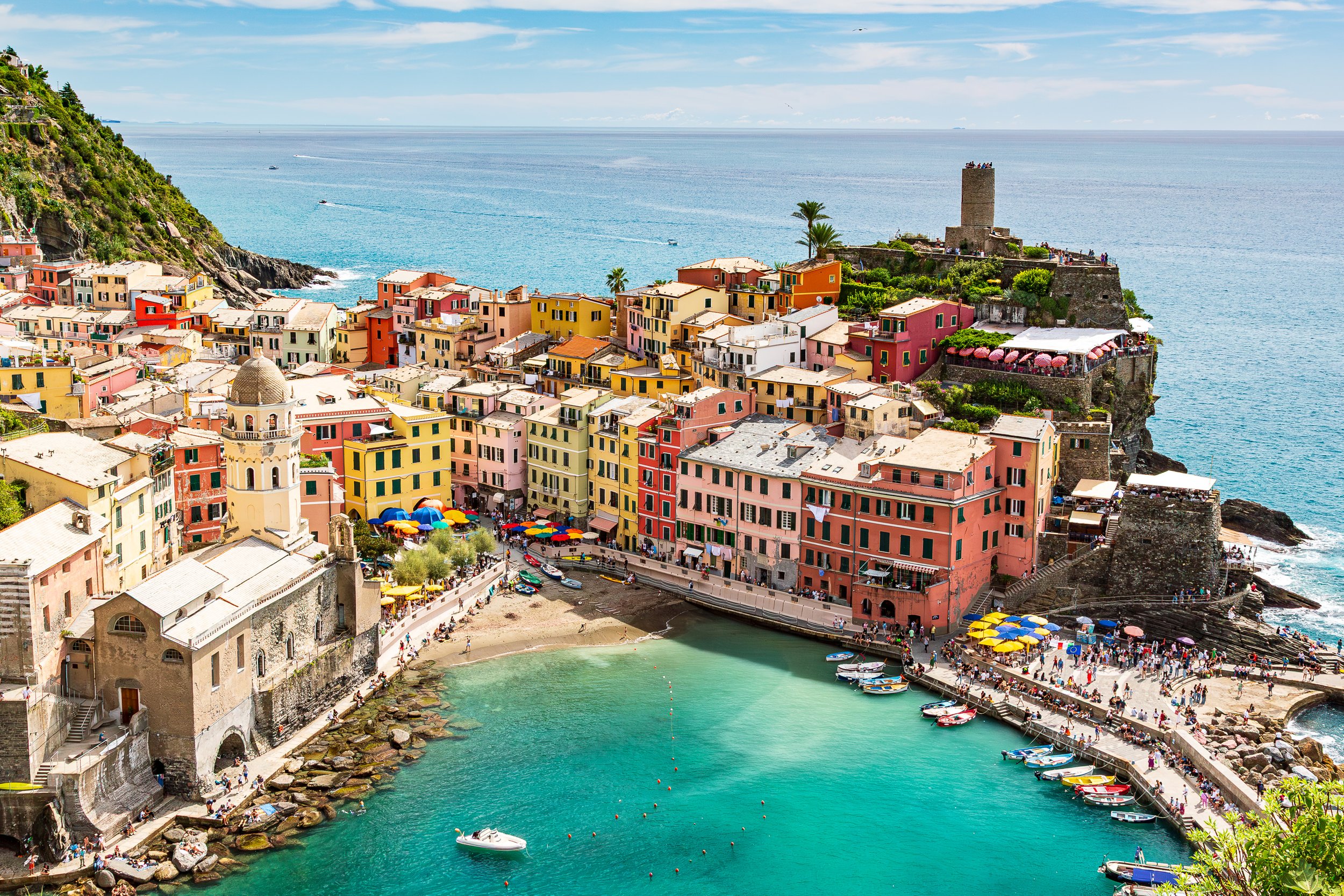 Colorful cliffside village with small boats in a harbor, beach, and walking crowd, overlooking the Mediterranean Sea.