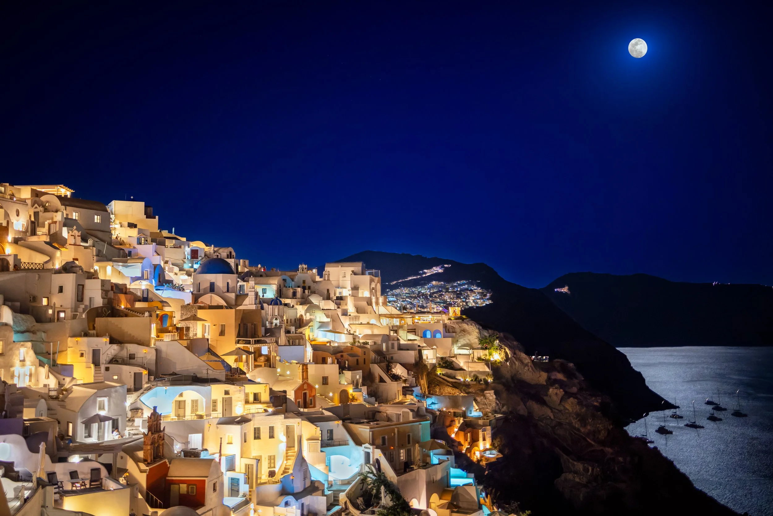 Nighttime view of white buildings on a hillside with multicolored lights, overlooking a dark sea under a moonlit sky.