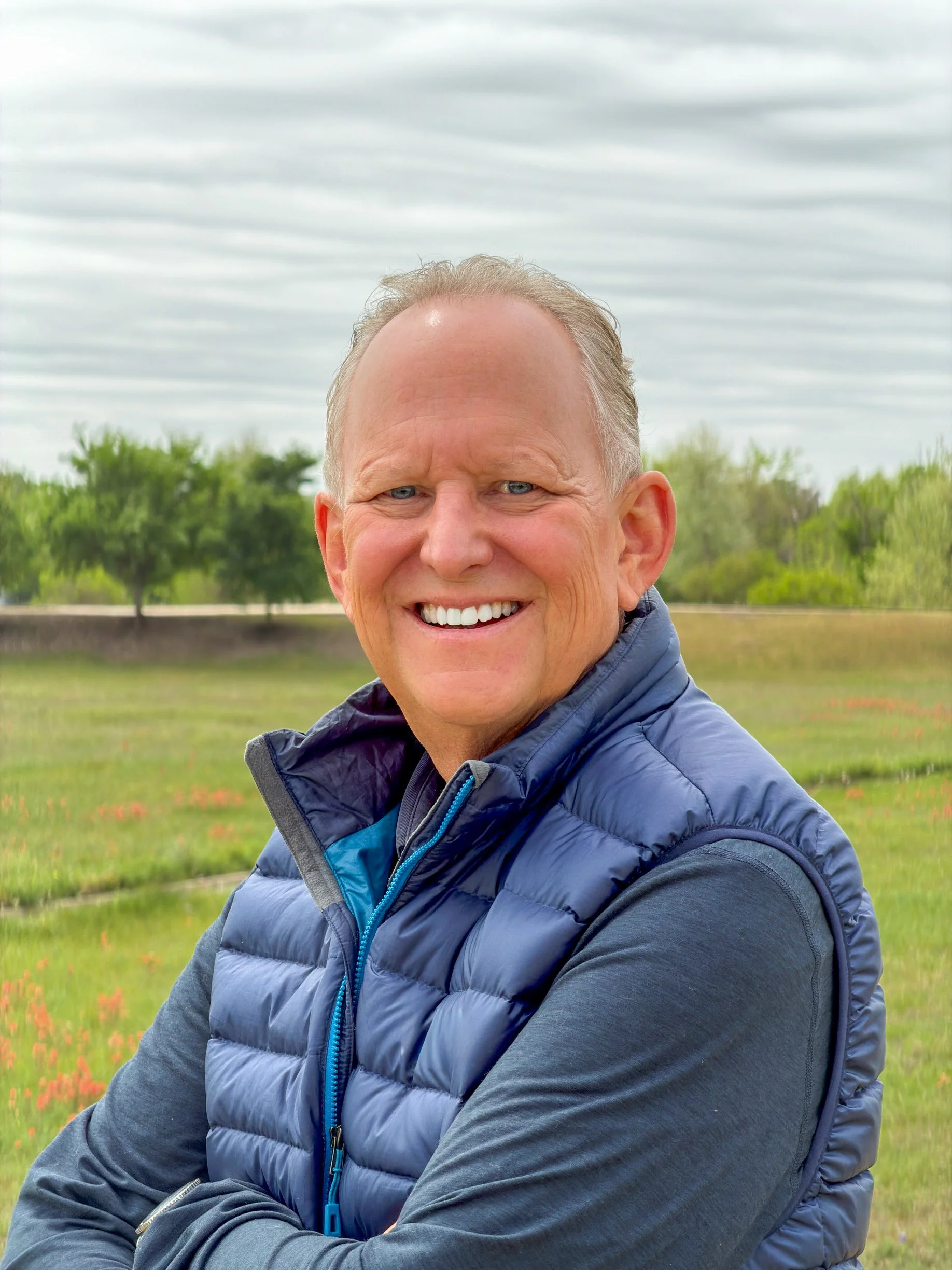 A smiling man outdoors wearing a blue puffer vest and gray long-sleeve shirt, with trees and cloudy sky in the background.