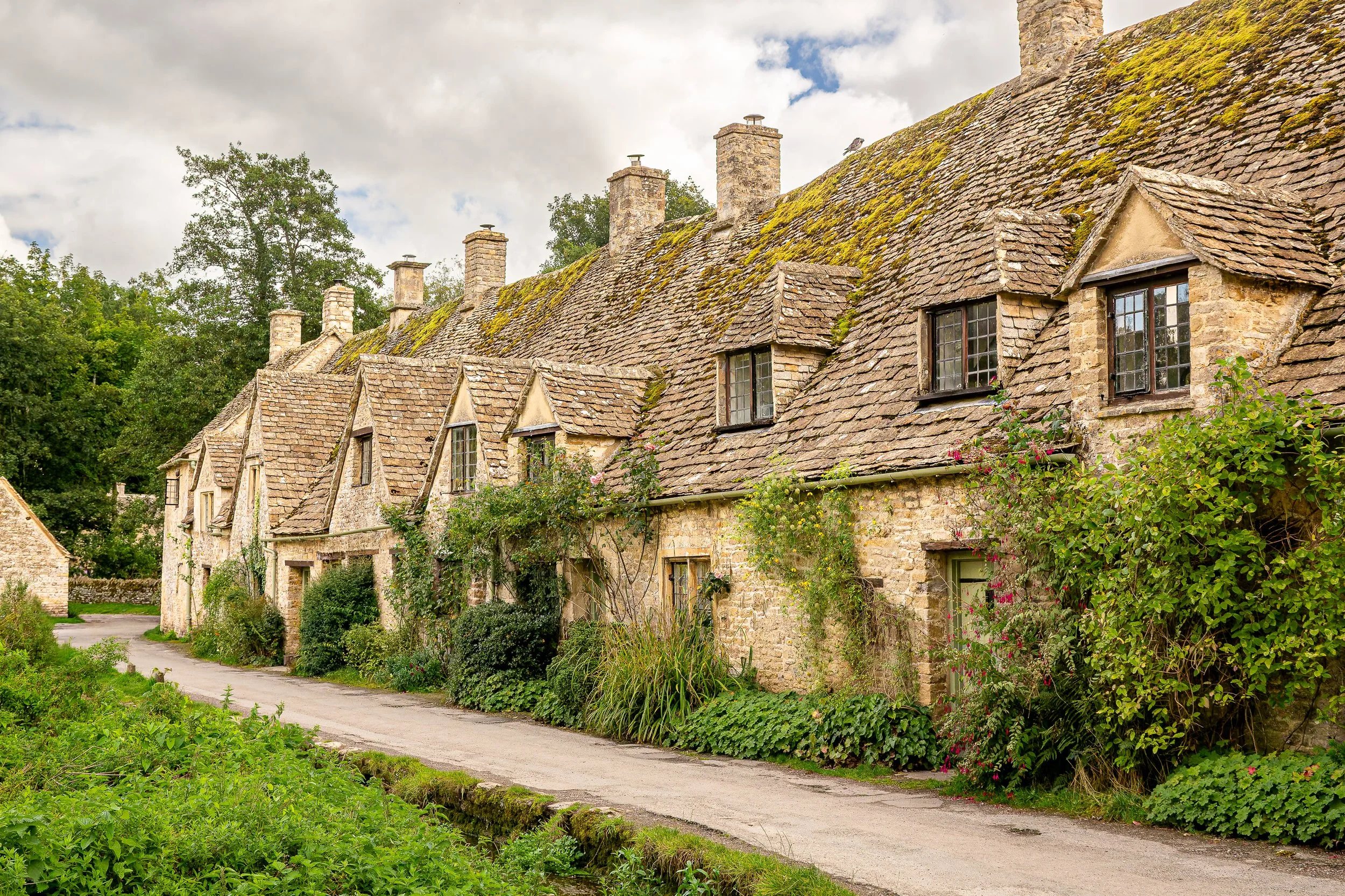 A row of old stone cottages with steep, moss-covered tile roofs, surrounded by lush greenery and flowering bushes on a narrow roadside, under a partly cloudy sky.