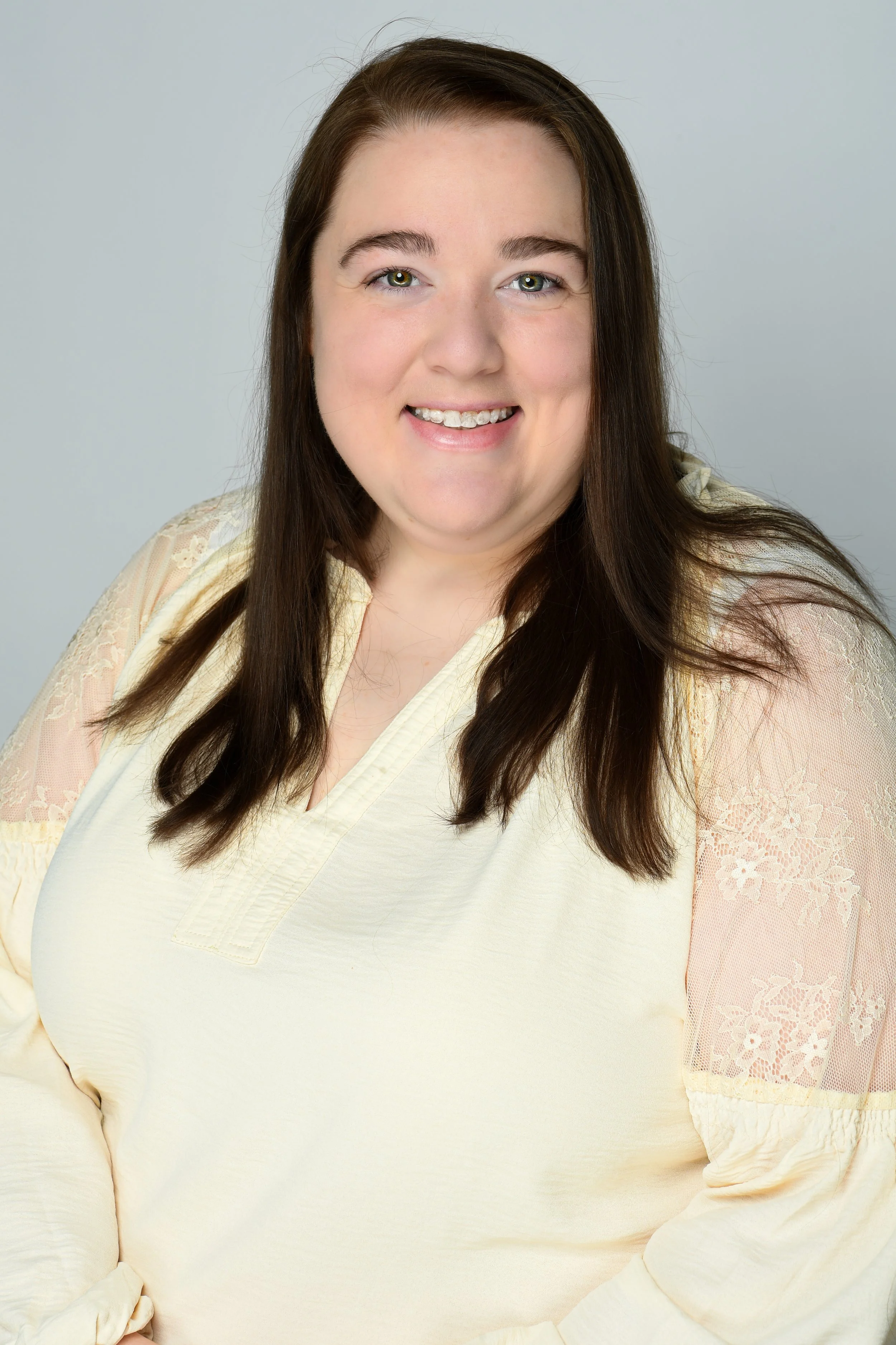 A young woman with long brown hair smiling, wearing a light yellow blouse with lace details on the sleeves, against a plain light gray background.