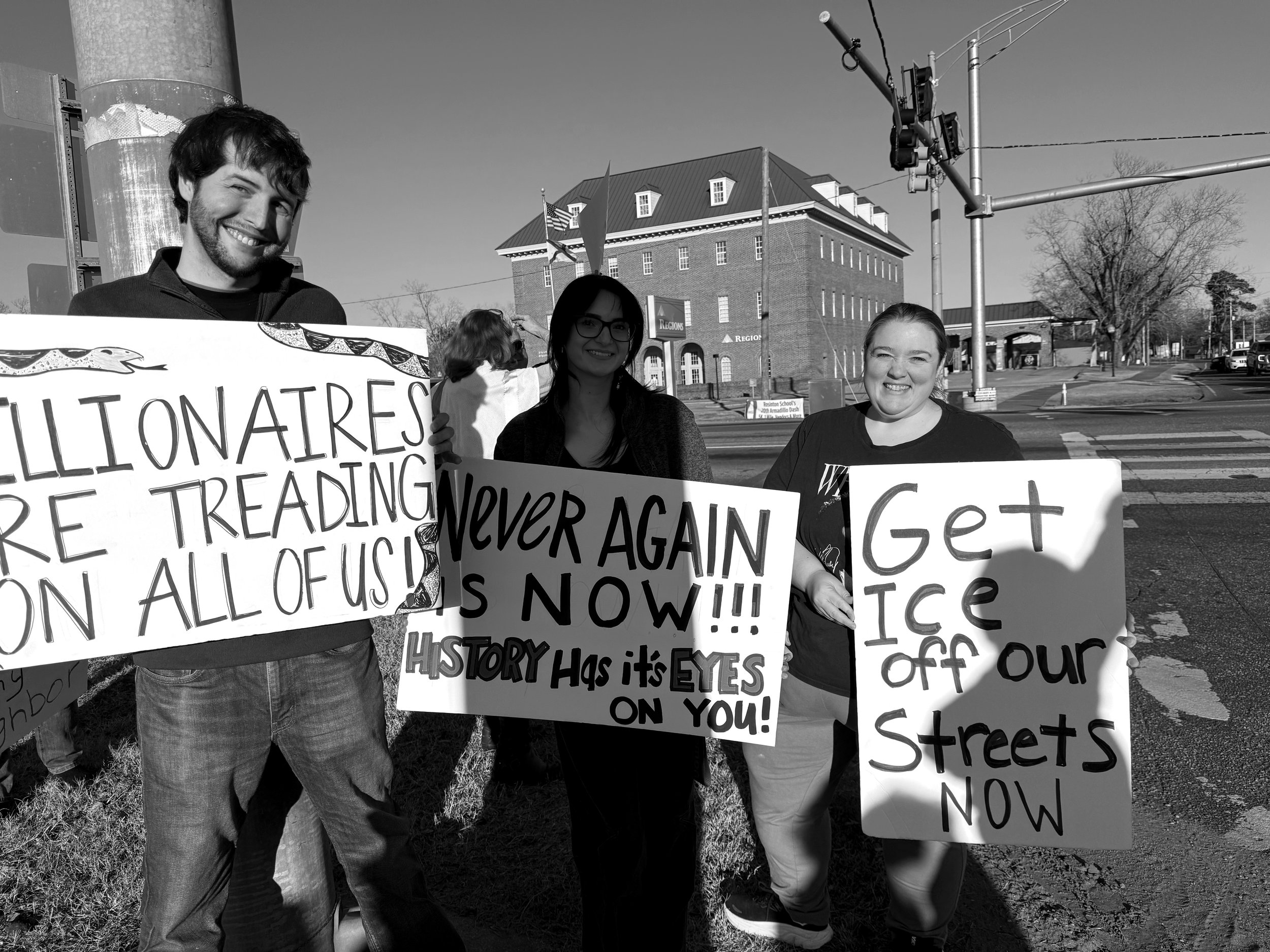 Three people protesting on a city street holding signs with hashtags and messages about social and political issues.