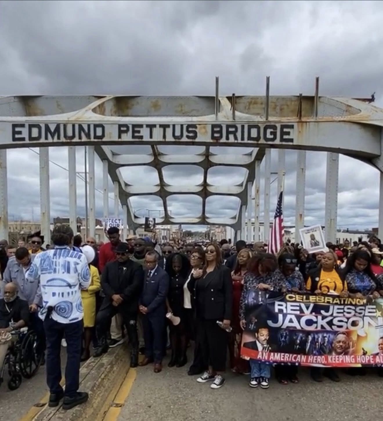 A large group of people gathered on a bridge with a sign reading 'Edmund Pettus Bridge.' Some are holding signs and a large banner celebrating Rev. Jesse Jackson. The sky is cloudy.