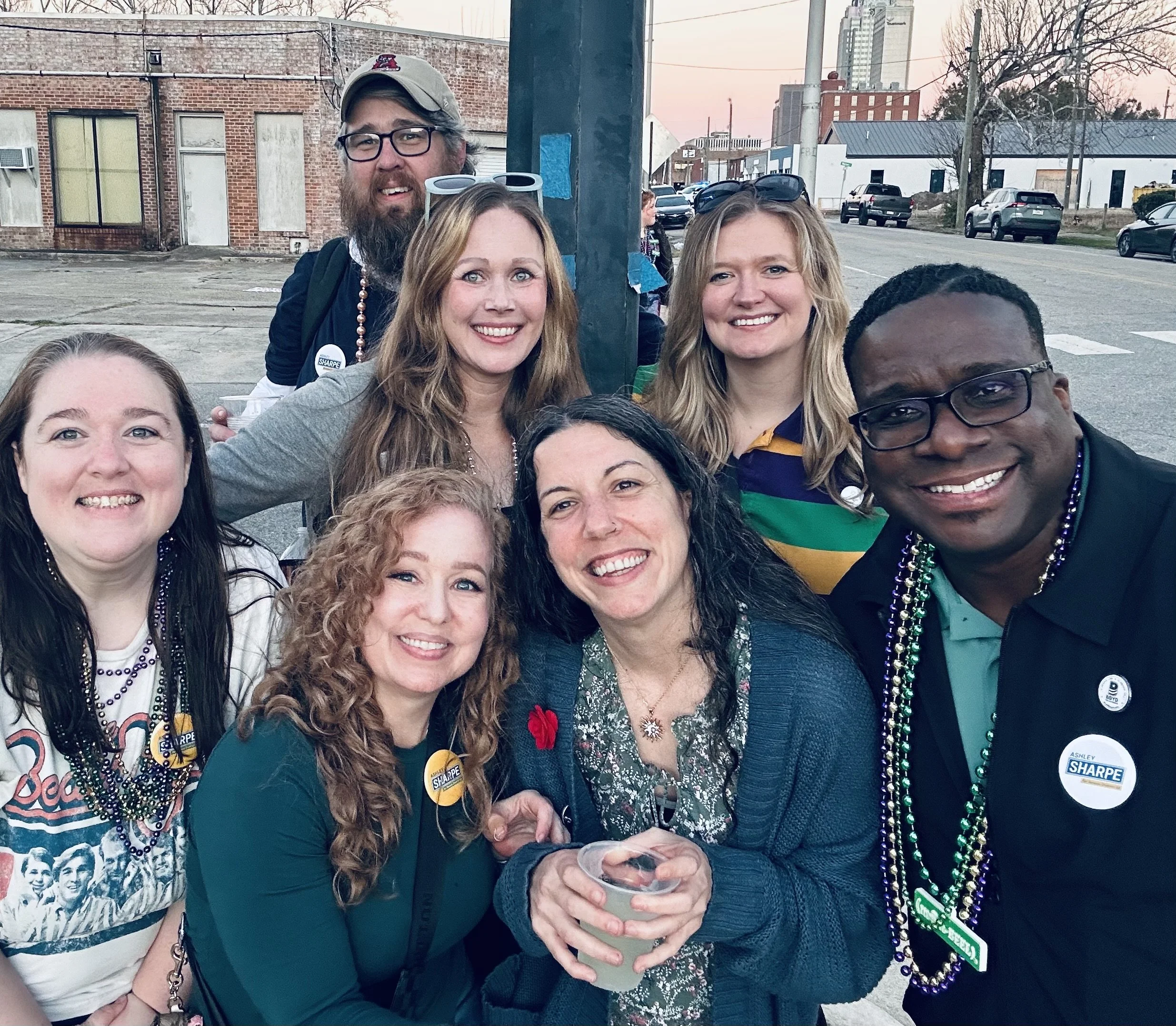 Group of friends smiling at an outdoor gathering during sunset, wearing campaign and Mardi Gras beads, with some holding drinks, in a city street setting.