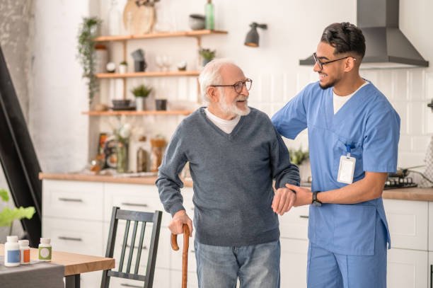 A healthcare worker smiling and supporting an elderly man with a cane in a bright, modern kitchen.