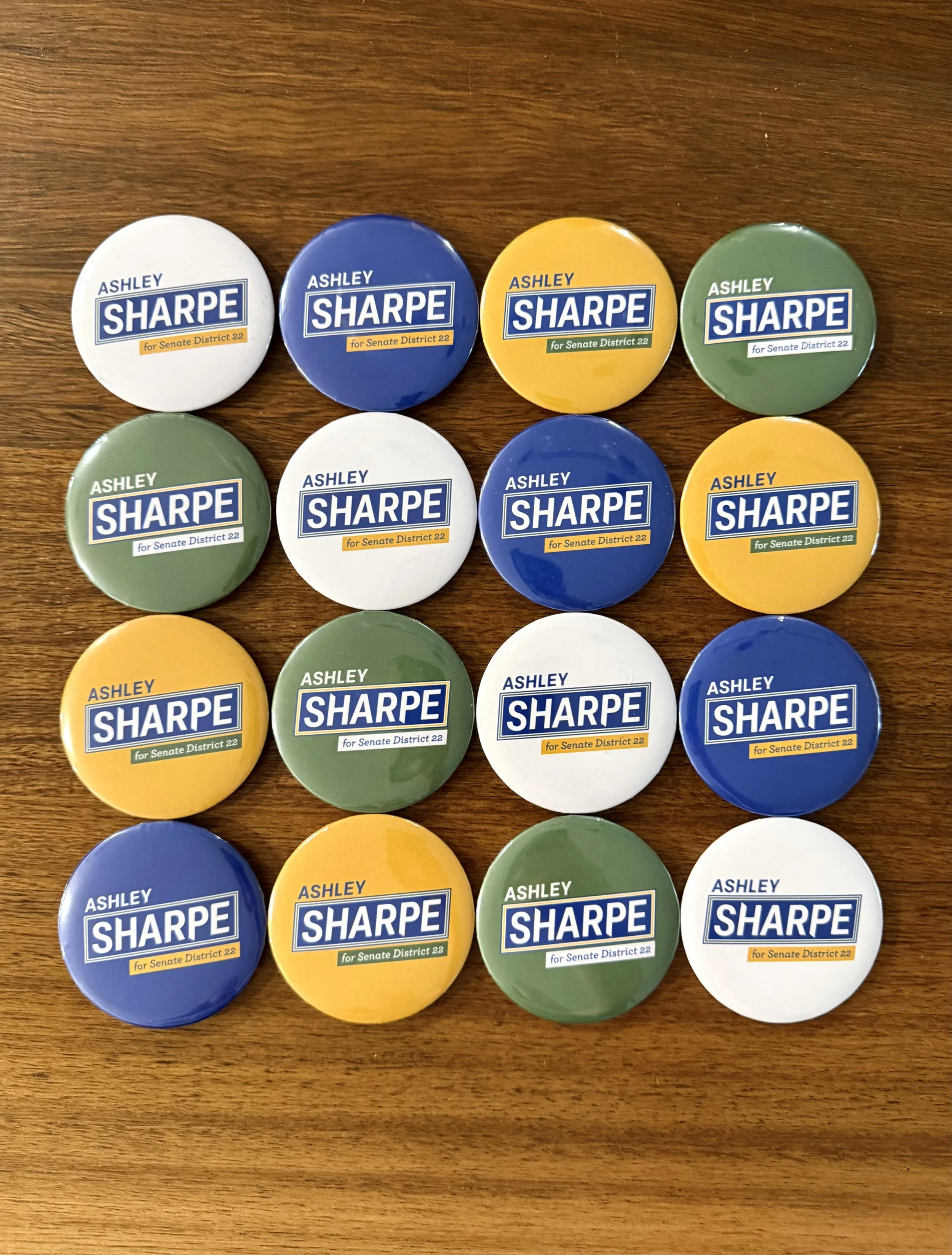 Pins featuring 'Ashley Sharpe for Senate District 22,' in white, blue, yellow, and green, arranged on a wooden surface.