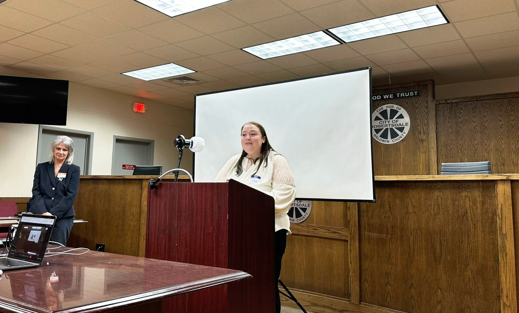 A woman standing at a wooden podium giving a speech in a conference room. Another woman stands to the side, near a table with a laptop. The room has a large white screen behind the speaker, wood-paneled walls, and ceiling lights. A sign on the wall indicates the location is in the city of Robertsondale, Baldwin County.