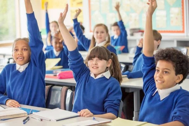 Elementary school classroom with students raising their hands, wearing blue uniforms.