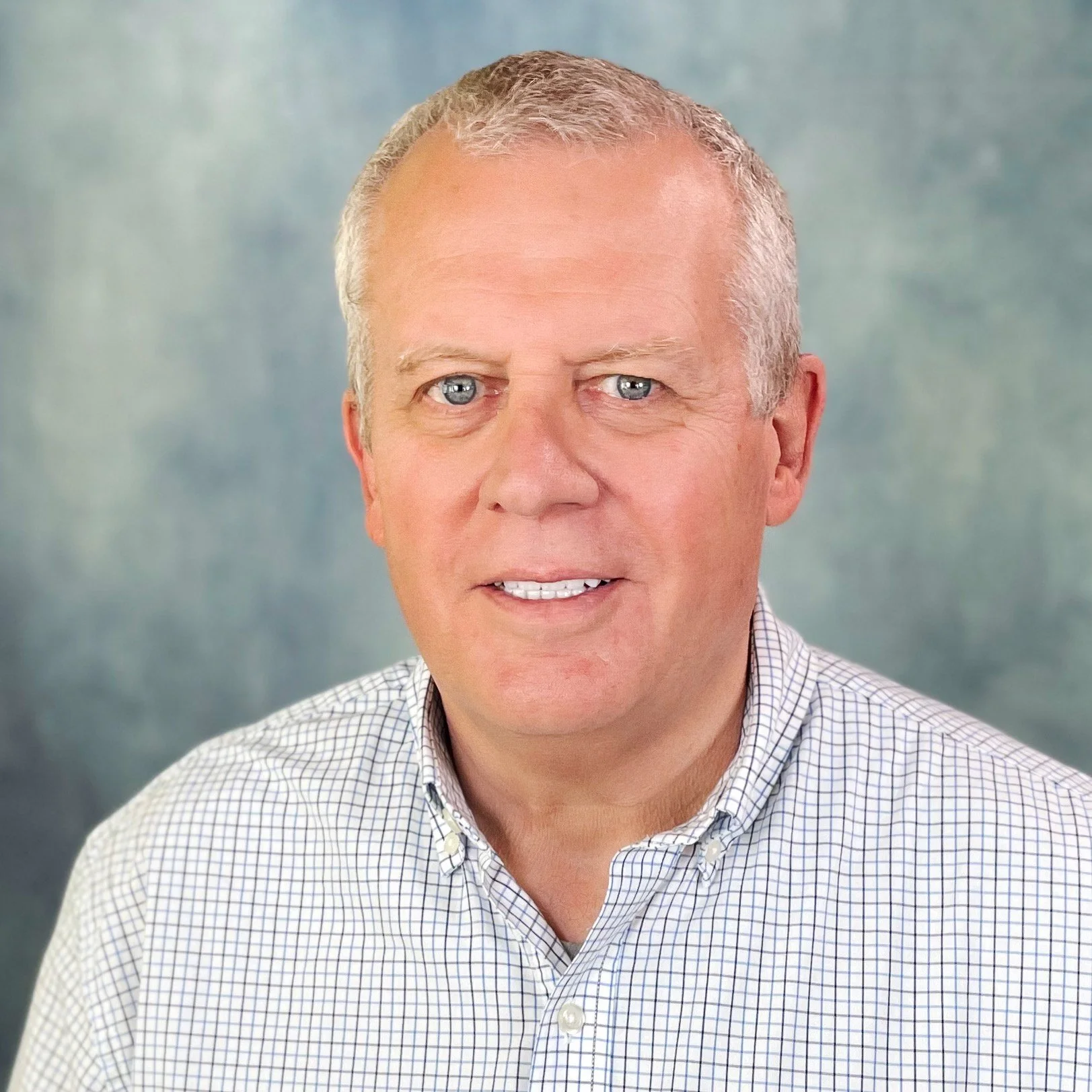 A middle-aged man with short gray hair and blue eyes, wearing a checkered white and blue shirt, smiling against a soft blue-gray background.