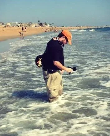 Man standing in shallow ocean water near the beach, wearing an orange cap, black hoodie, and beige shorts, holding a field recording equipment.