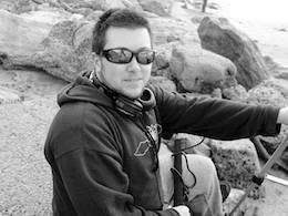 Man in sunglasses wearing a hoodie, sitting outdoors near rocks, holding a device.