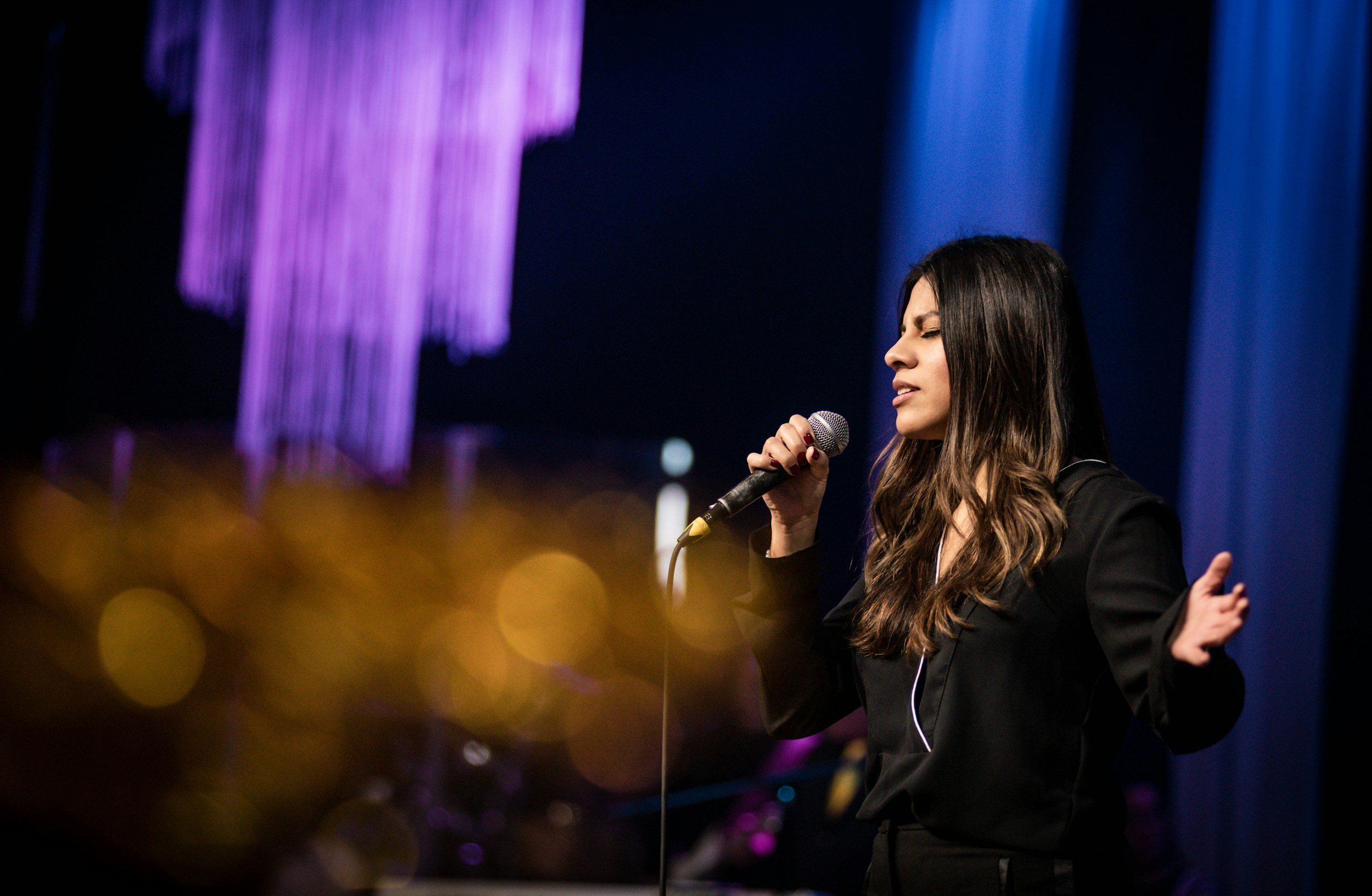 A woman singing into a microphone on stage with purple and blue lighting in the background.