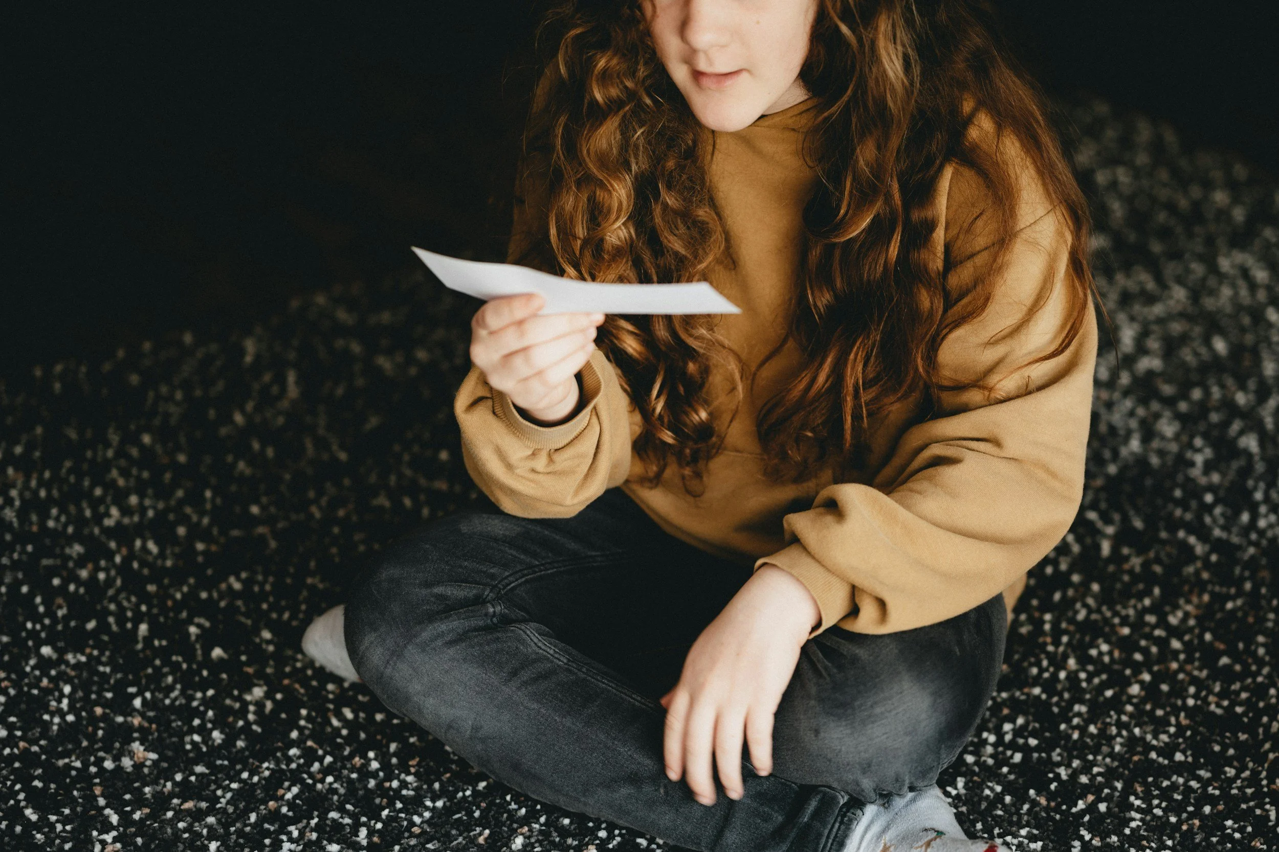 A young woman with long curly red hair sitting cross-legged on a black and white speckled carpet, looking at a piece of paper in her hand.