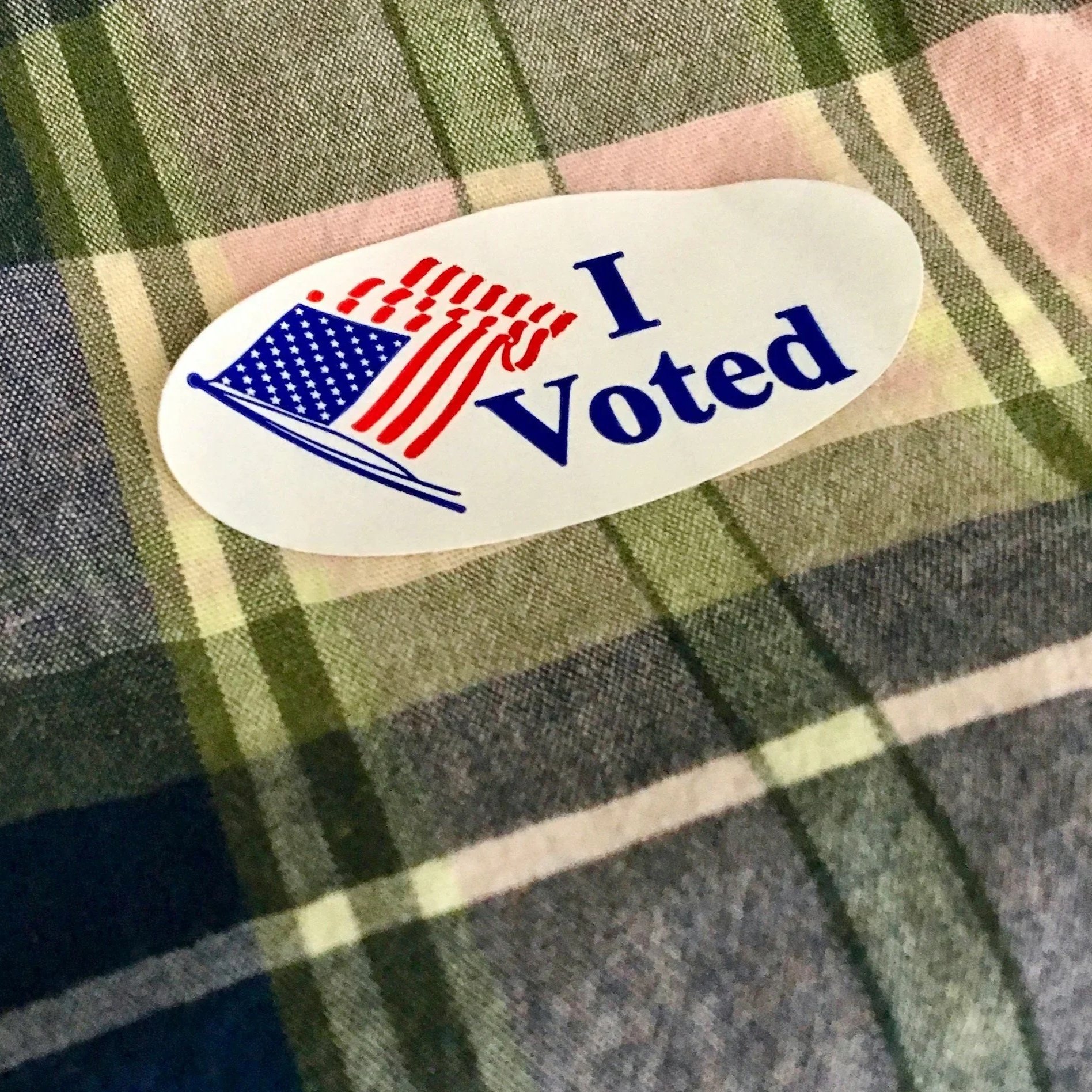 An oval sticker with an American flag and the words "I Voted" on a plaid fabric surface.