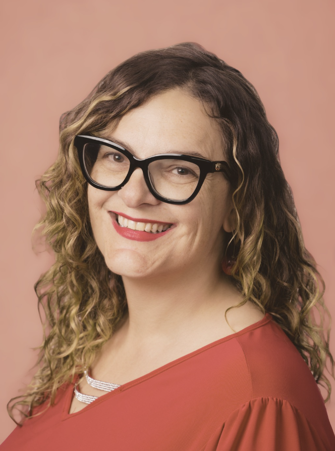 A woman with curly hair, wearing glasses and a red top, smiling against a pink background.