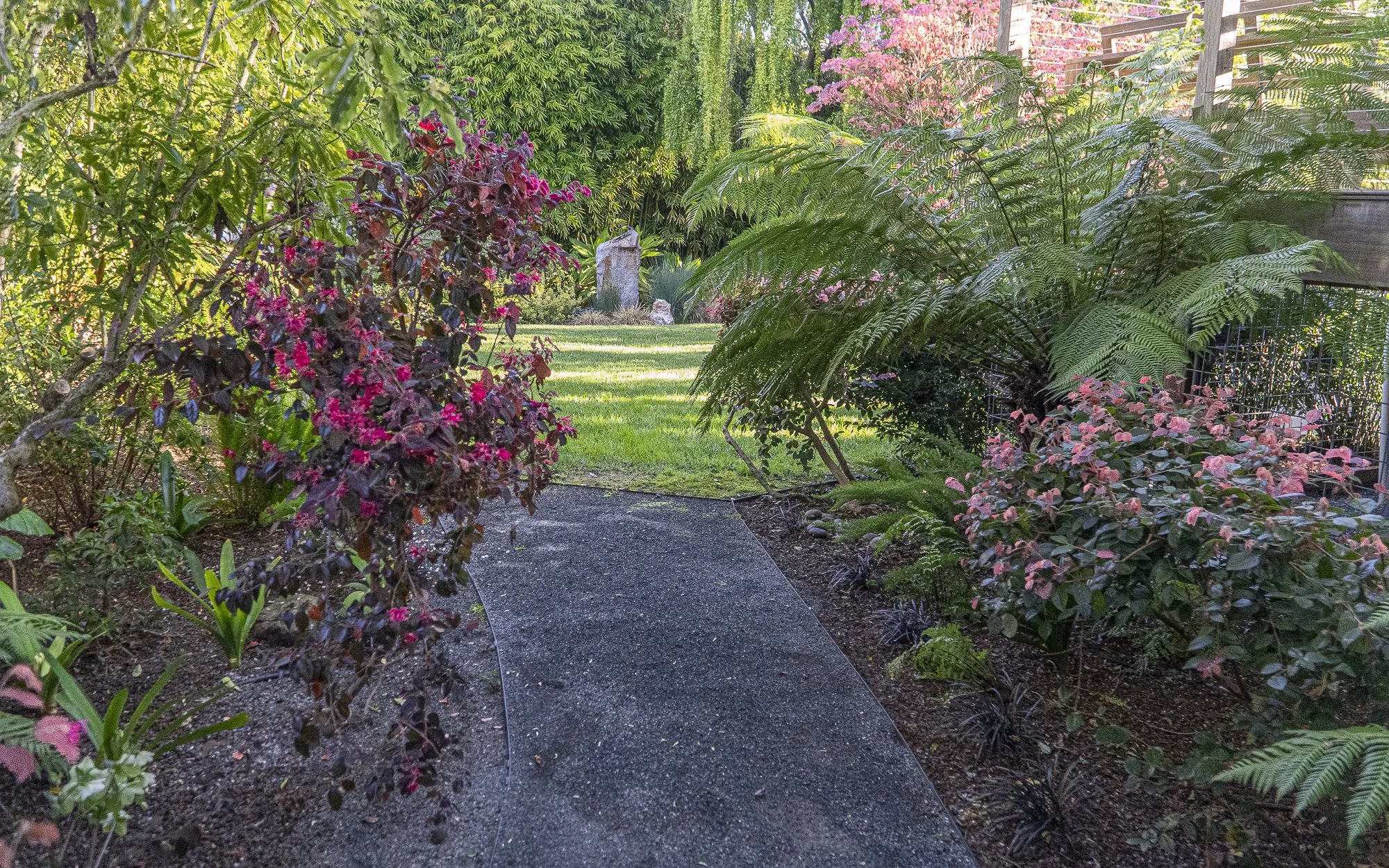 A garden pathway surrounded by colorful flowering plants and lush greenery, leading to an open grassy area with flower sculptures in the background.