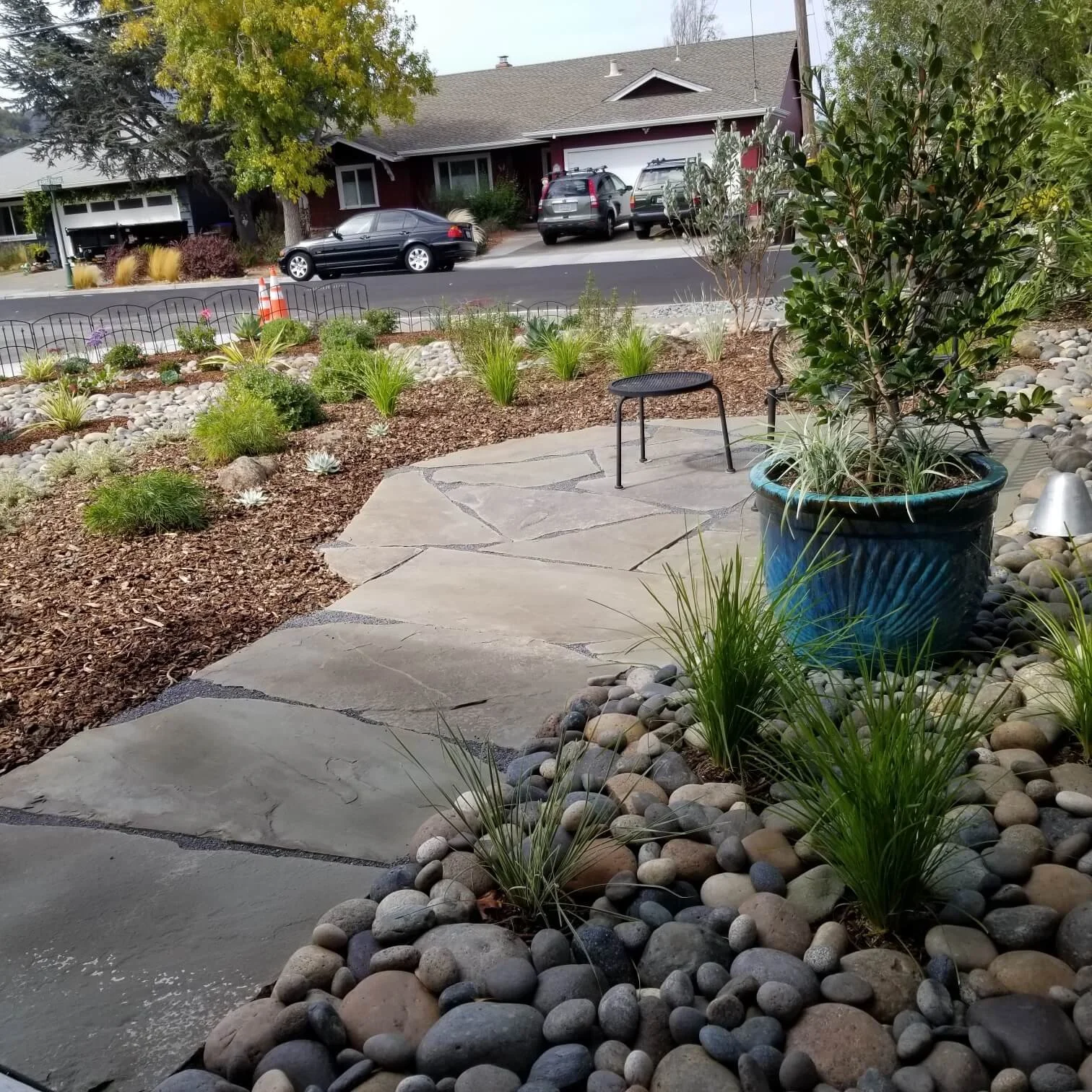 A front yard with a pathway made of large pavers, surrounded by decorative rocks and various small plants and shrubs. There is a large blue pot with a shrub and other plants, and a black stool nearby. In the background, a street with parked cars and a house across the street with trees and plants around it.