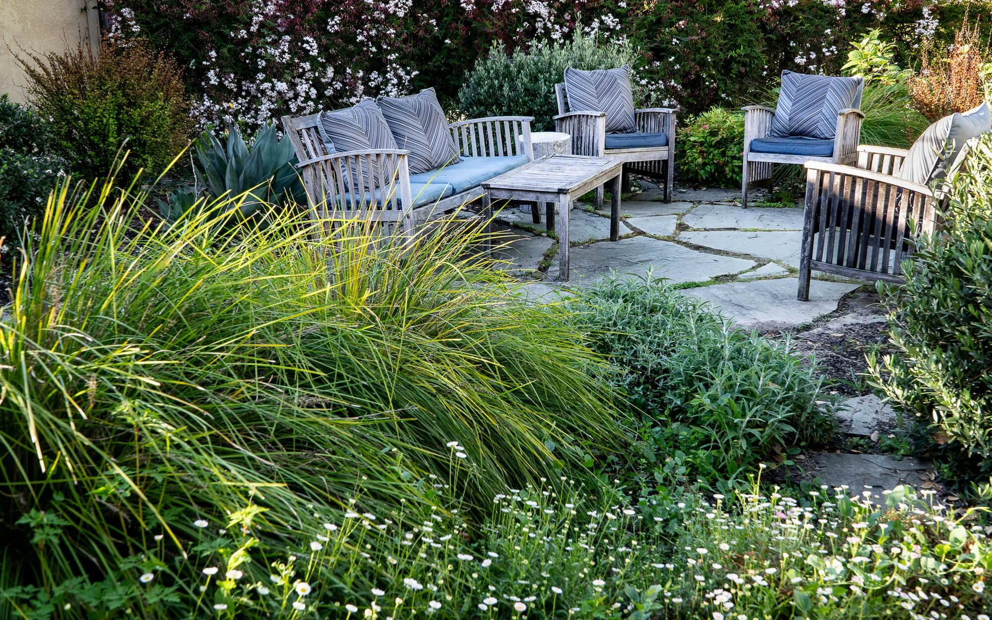 Stone pathway with outdoor wooden seating area surrounded by lush greenery and flowers.