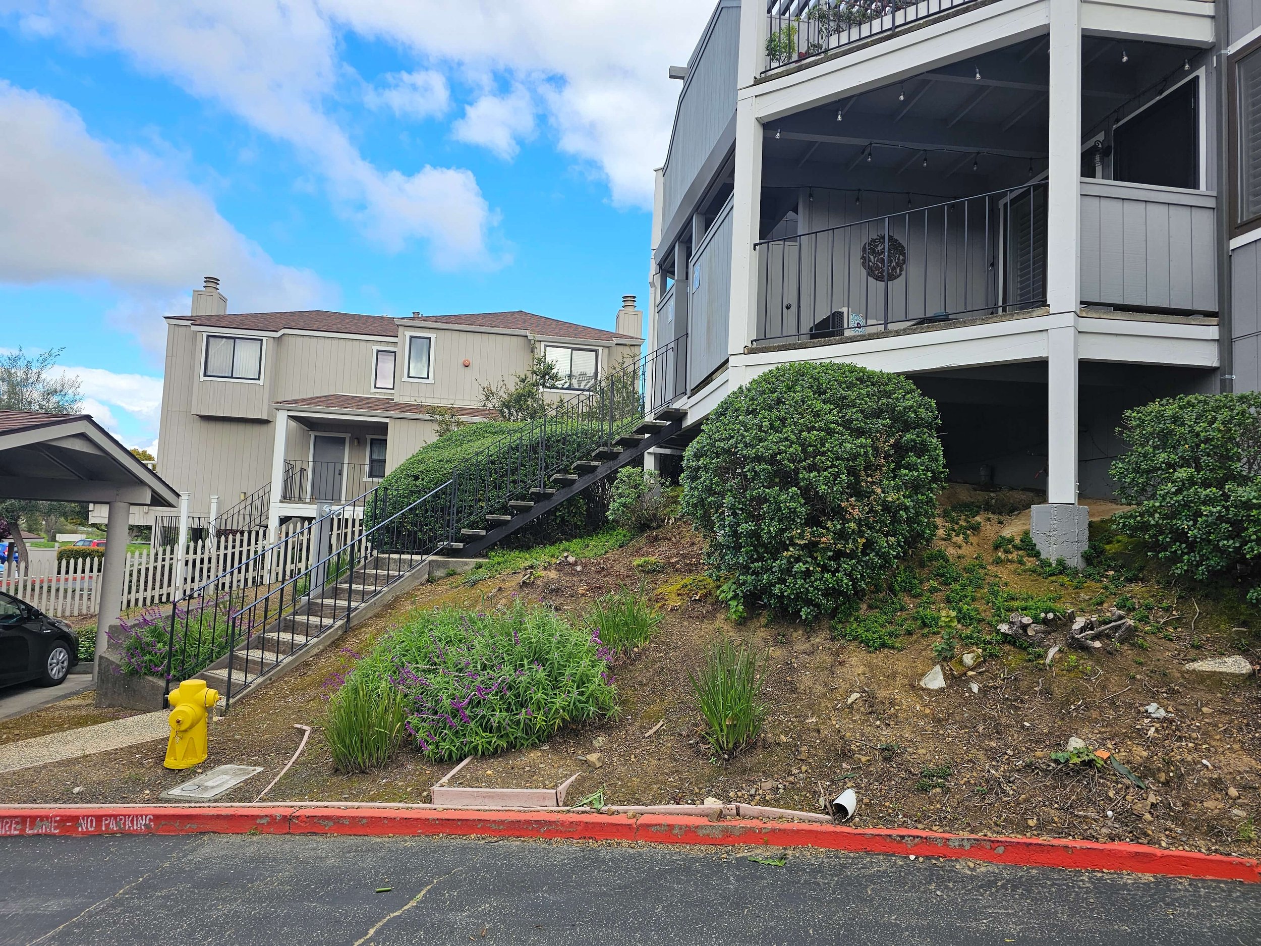 Multi-story residential buildings with balconies, stairs, and greenery in a suburban setting on a partly cloudy day.