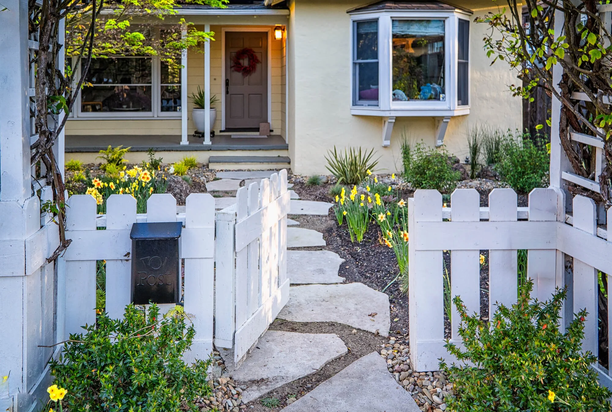 Front yard garden with stepping stones, yellow flowers, green bushes, white picket fence, and yellow house with front porch and bay window.