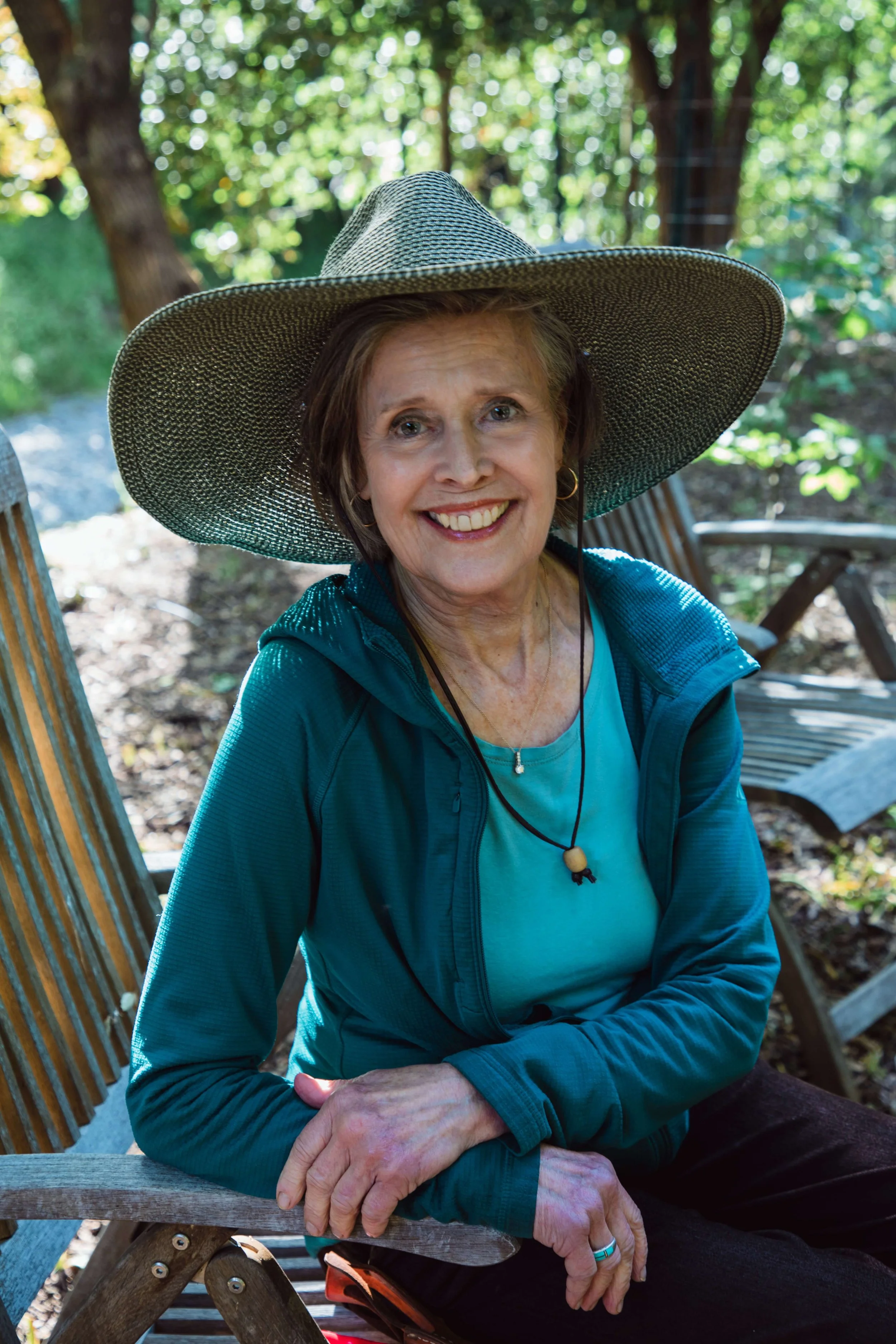 An older woman with short brown hair smiles while sitting outdoors on a wooden bench. She wears a wide-brimmed straw hat, a teal zip-up jacket, and earrings with a striped necklace. There is greenery and trees in the background.