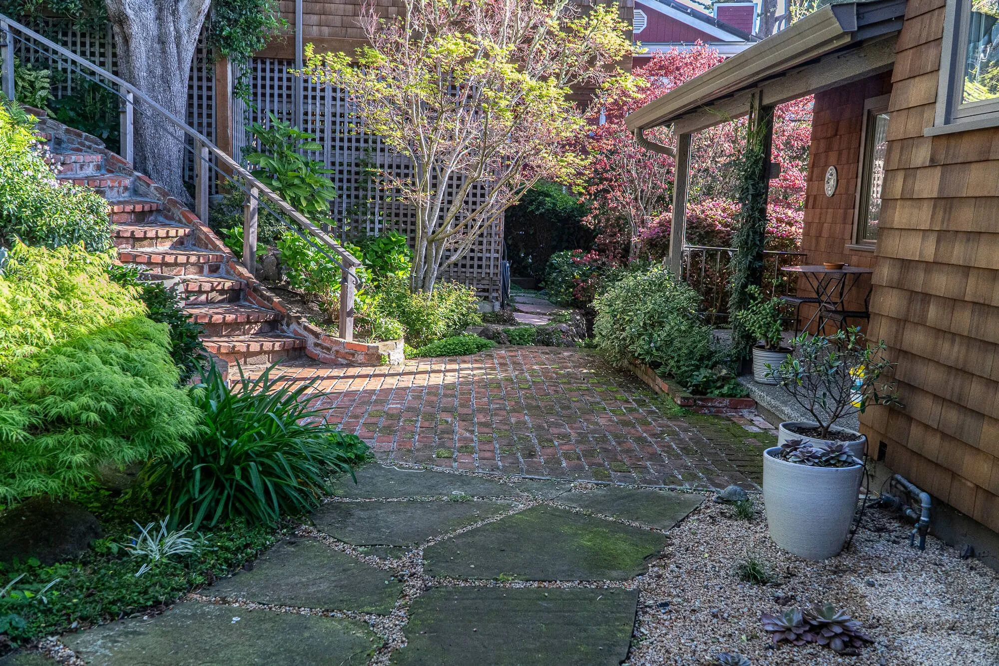 A cozy backyard with brick and stone pathway, wooden stairs, lush green plants, and pink flowering trees creating a peaceful garden setting.