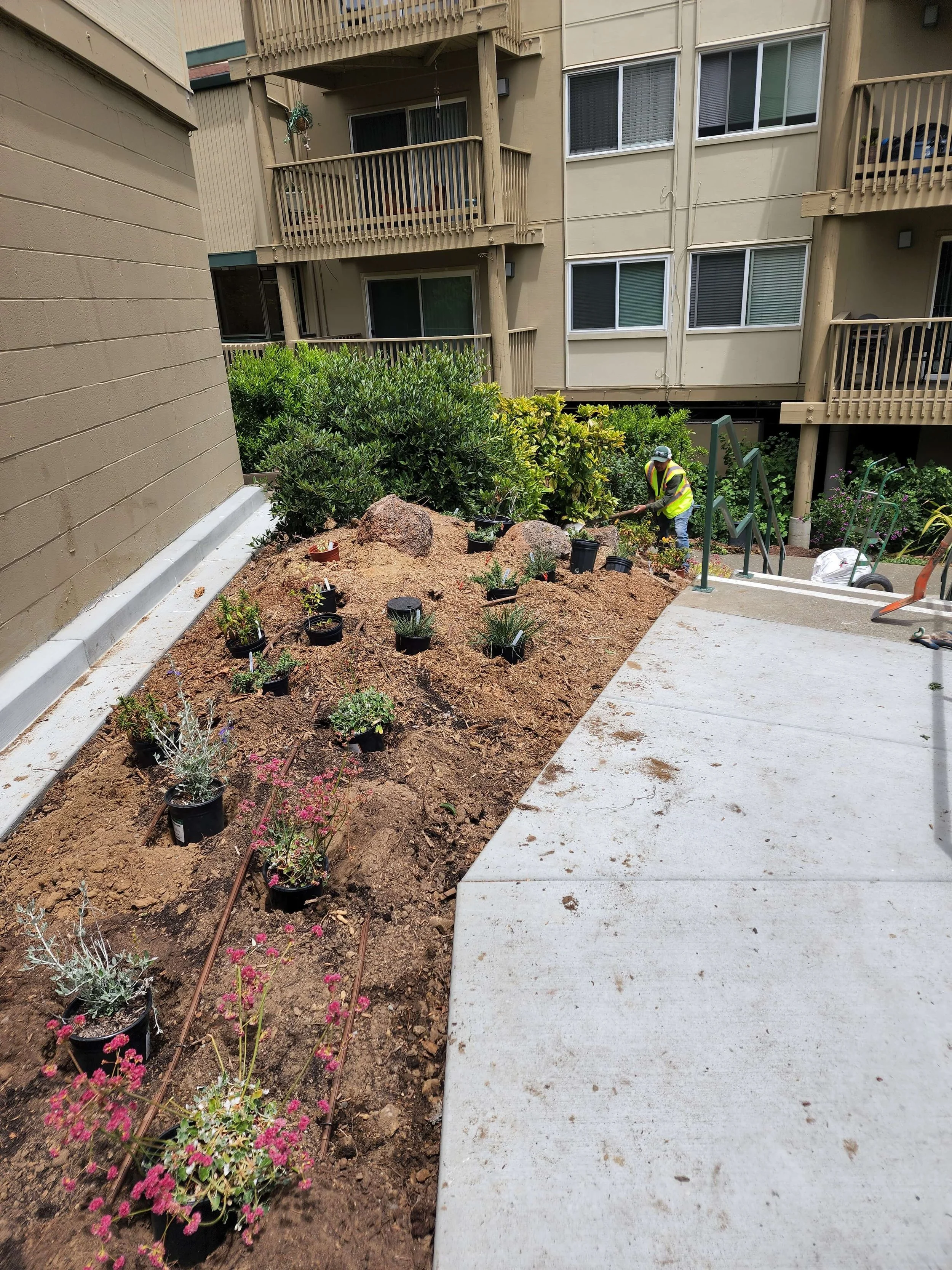 A construction worker in a yellow safety vest and helmet planting flowers in a garden bed beside a concrete sidewalk, with apartment buildings in the background.