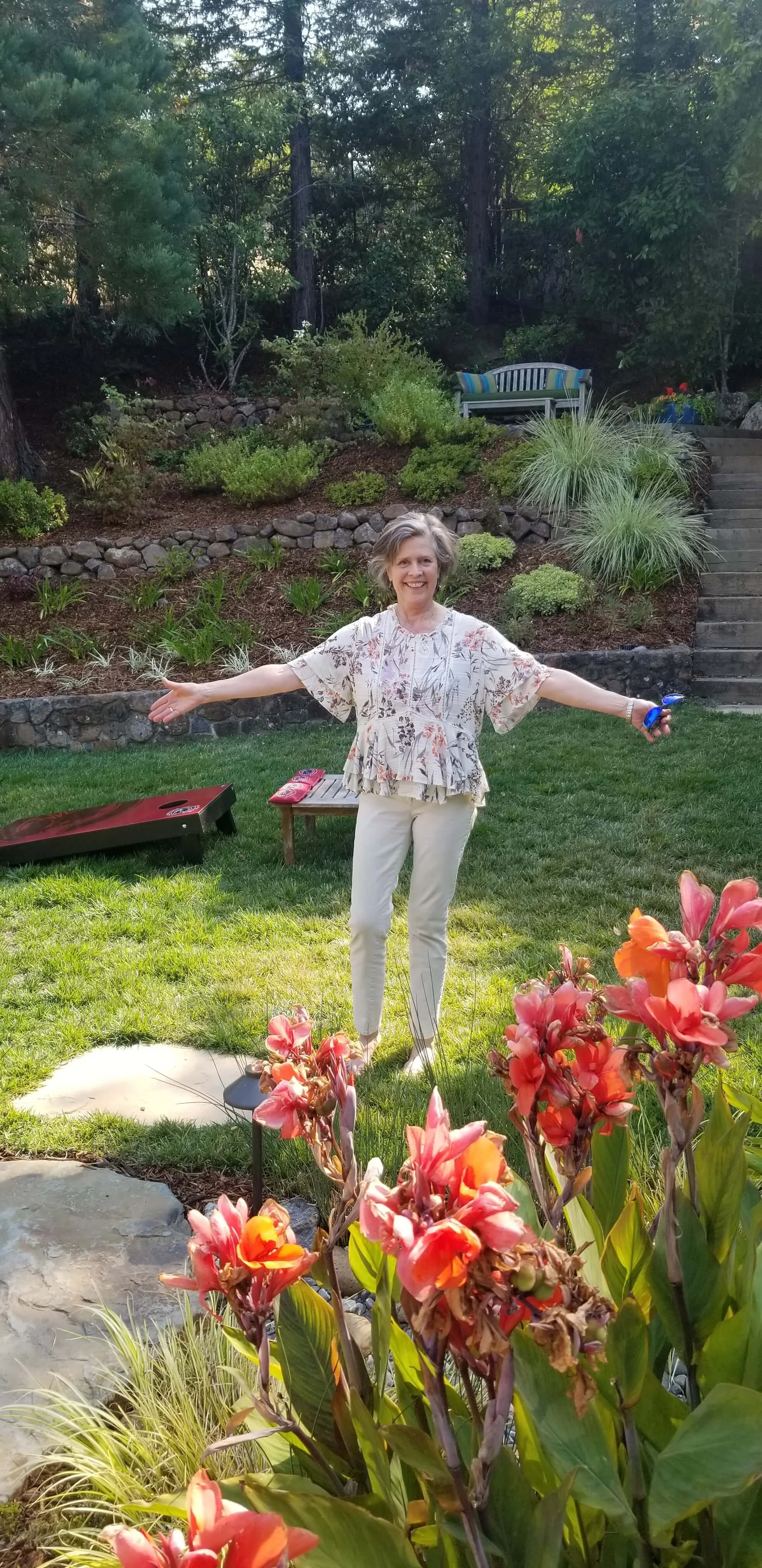 A woman standing in a lush backyard garden with her arms outstretched and smiling. She is wearing a floral blouse and light-colored pants, with colorful flowers and greenery around her.