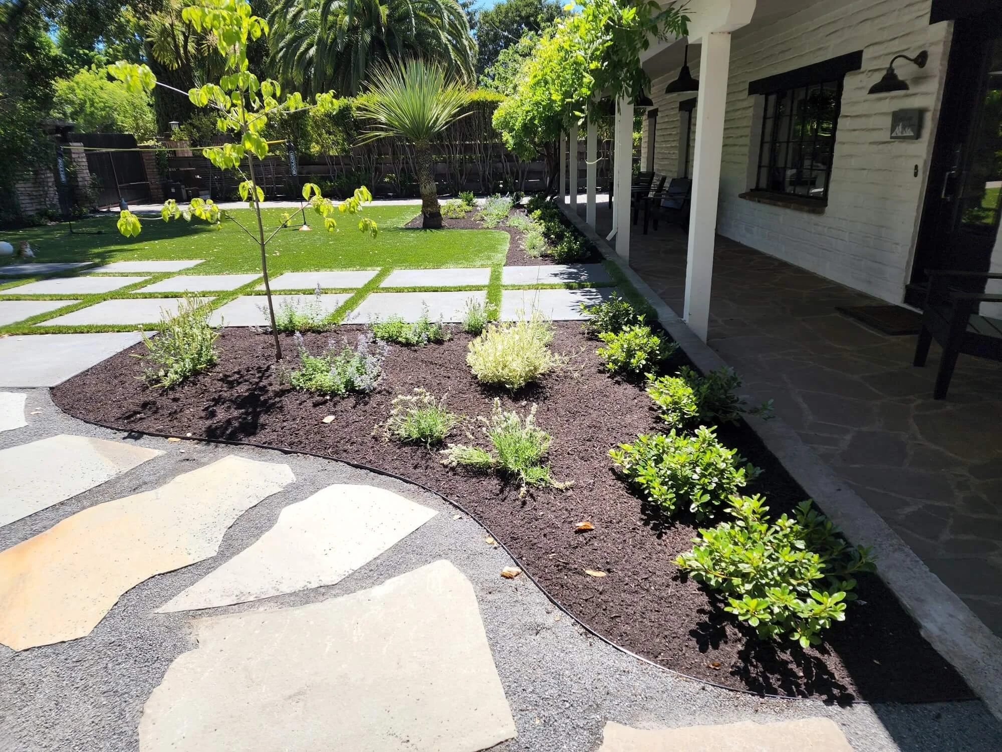 A landscaped backyard with a patio, a small tree, and various plants and bushes along a garden bed. The yard has a grassy area with stepping stones, and a house with a porch on the right side.
