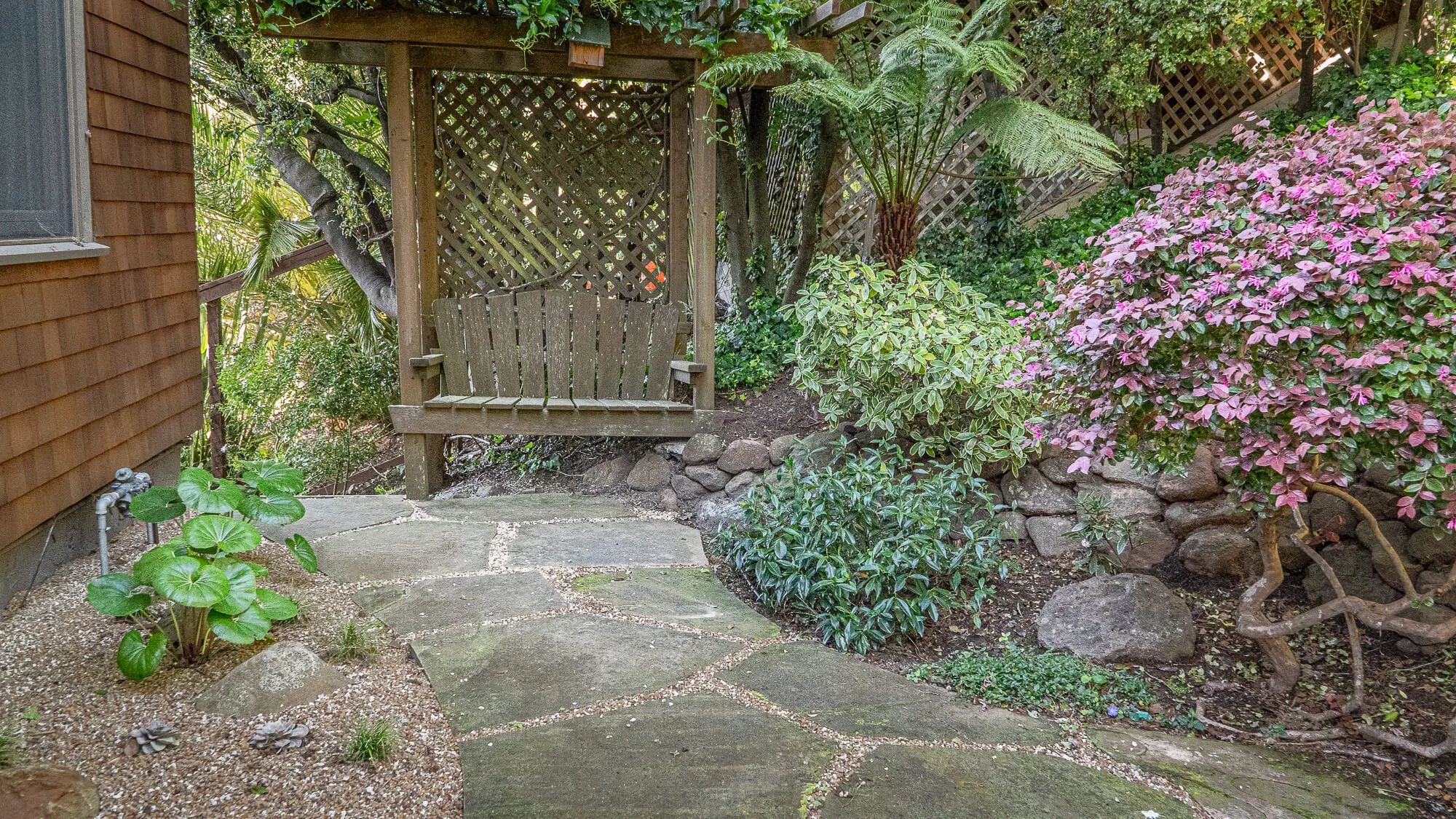 A small garden sitting area with a wooden bench swing under a wooden gazebo. The garden has various green plants and colorful pink flowering bushes, with a stone pathway leading to the swing.