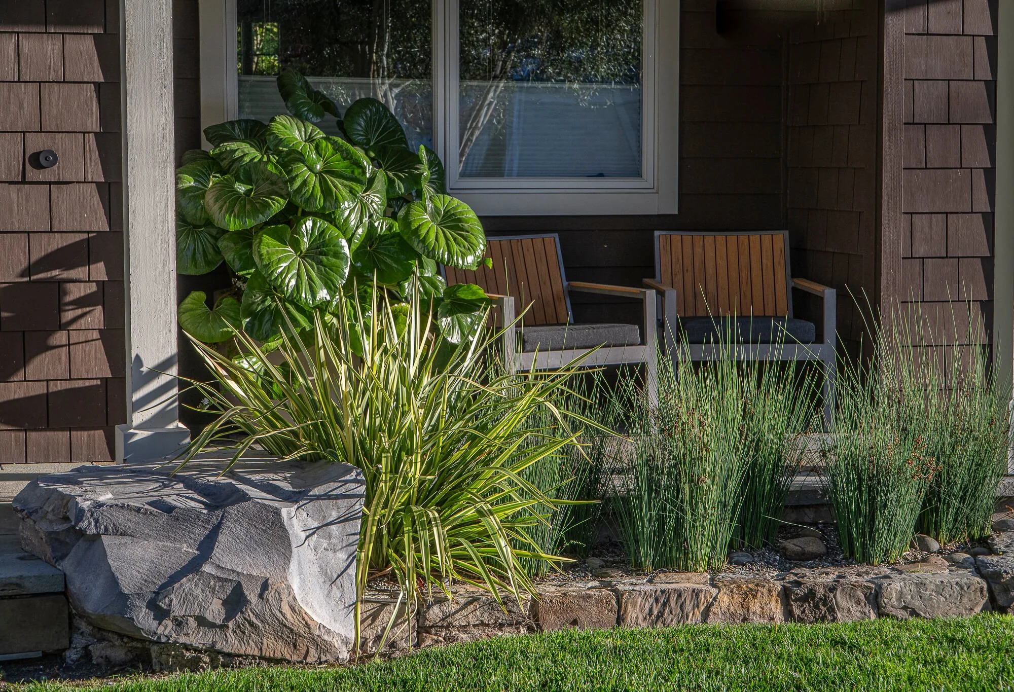 Front porch with two wooden chairs, large leafy green plants, and ornamental grasses in a landscaped garden bed with rocks.