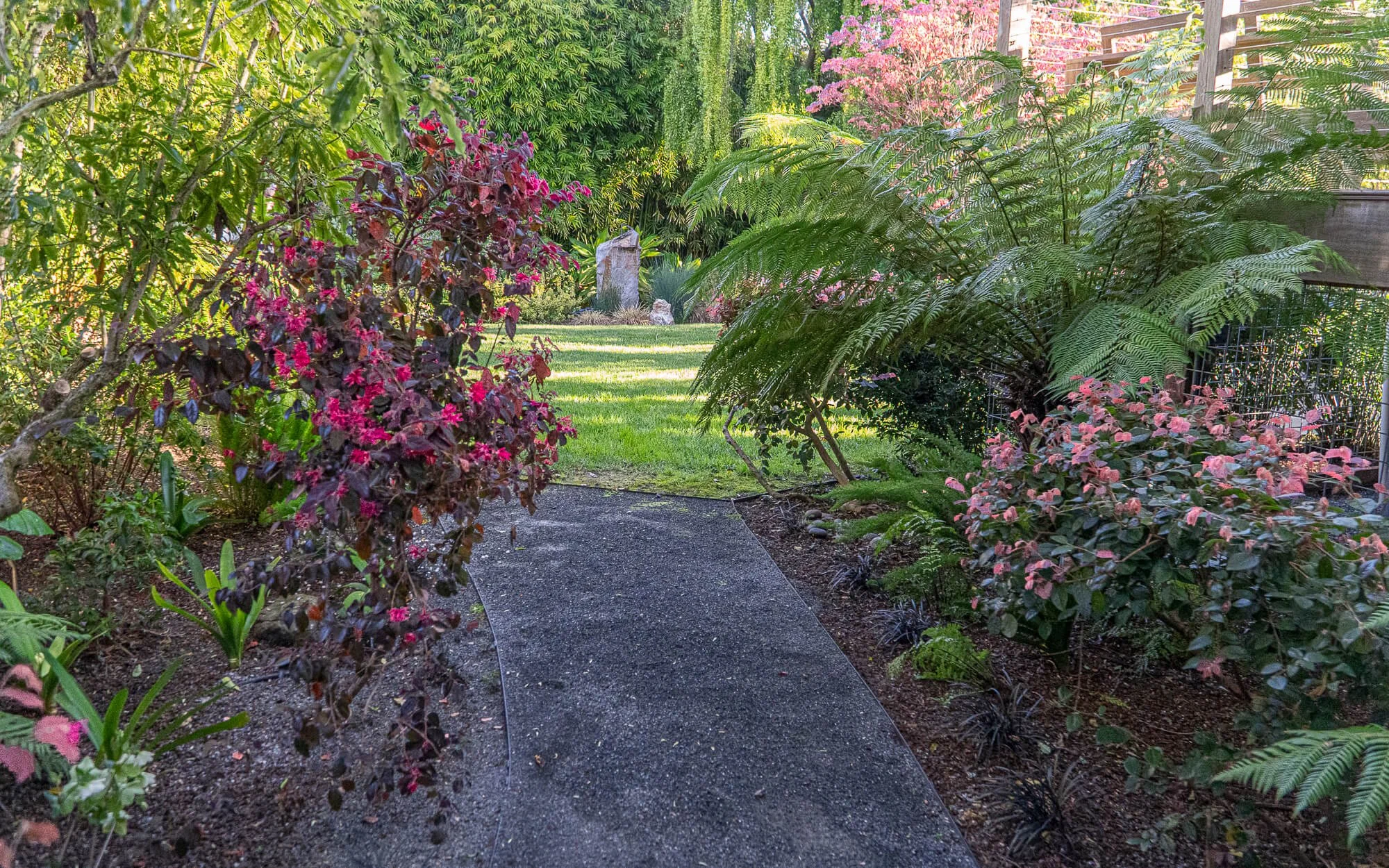 A lush garden pathway with colorful bushes and green foliage, leading to a bright open grassy area with trees and a few decorative stones in the background.