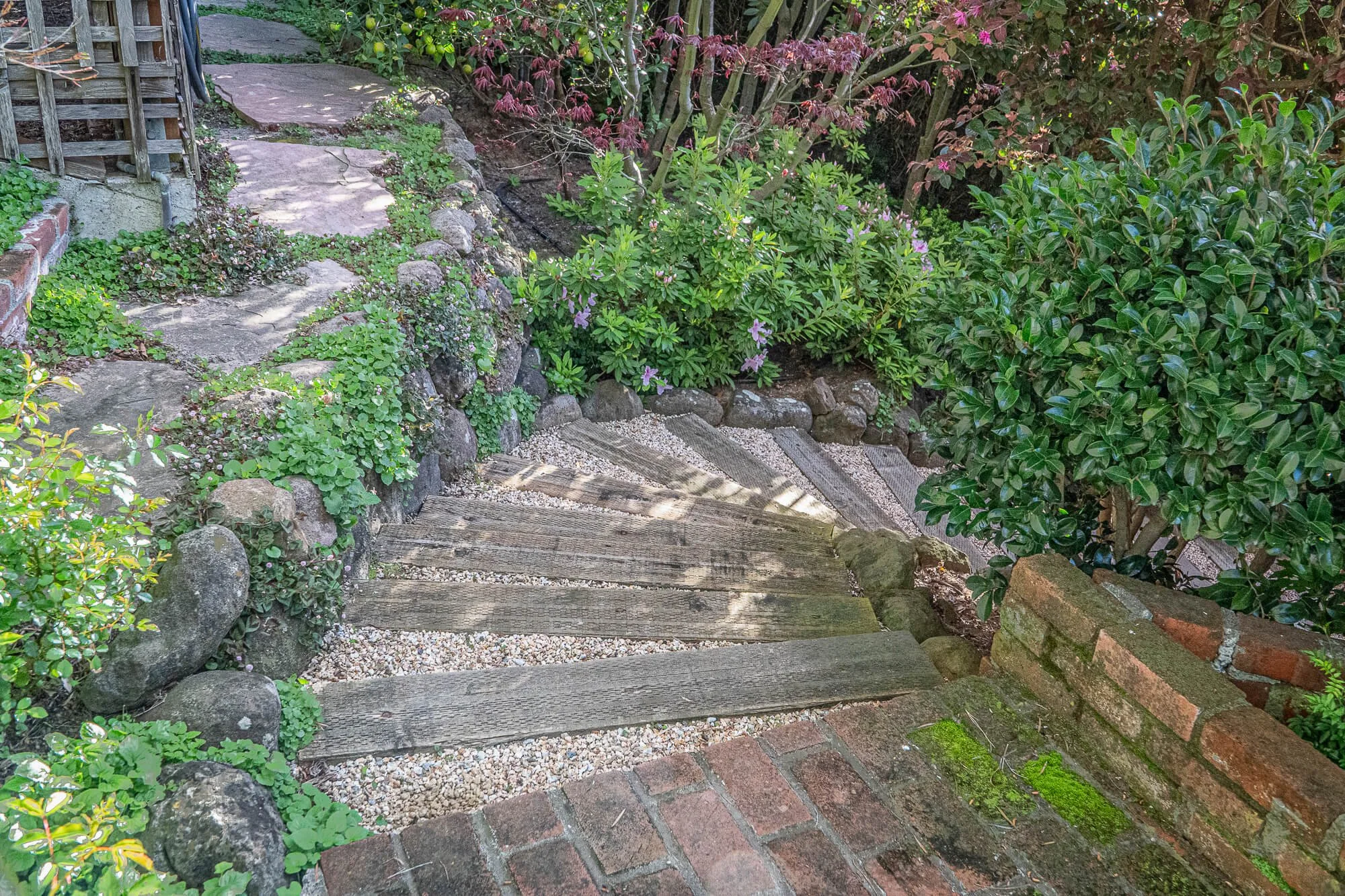 A rustic garden pathway with wooden steps bordered by rocks, gravel, and lush green plants and shrubs, leading up a small slope.