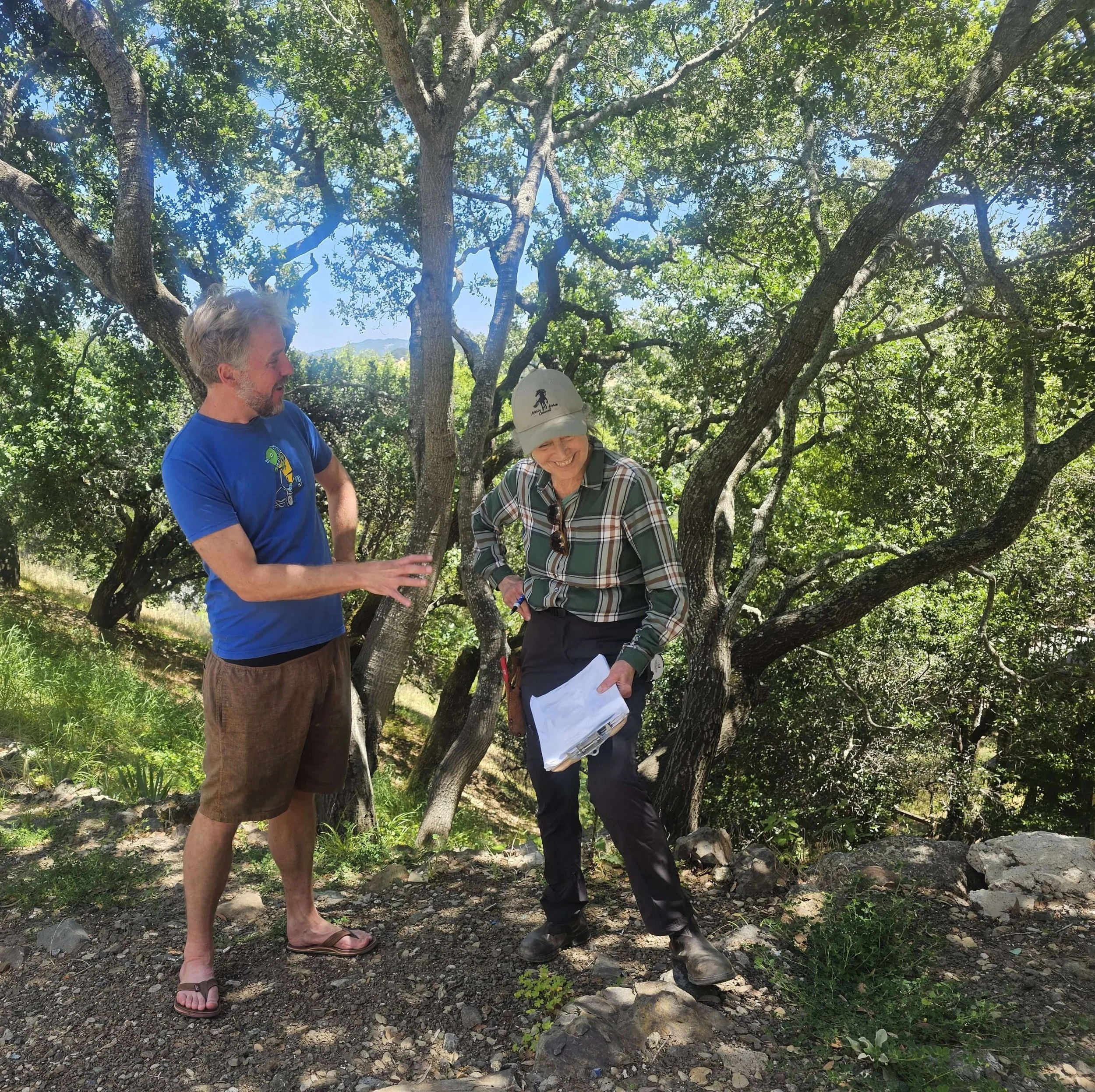 Three people are standing outdoors under trees on a sunny day. One person, a man, is wearing a blue t-shirt with a graphic and brown shorts, and flip-flops. He has gray hair and a beard. A woman, wearing a baseball cap, plaid shirt, and sunglasses, is looking down at some papers or maps, smiling. The third person is partially visible, wearing dark clothing and a hat, and appears to be holding a hiking pole.