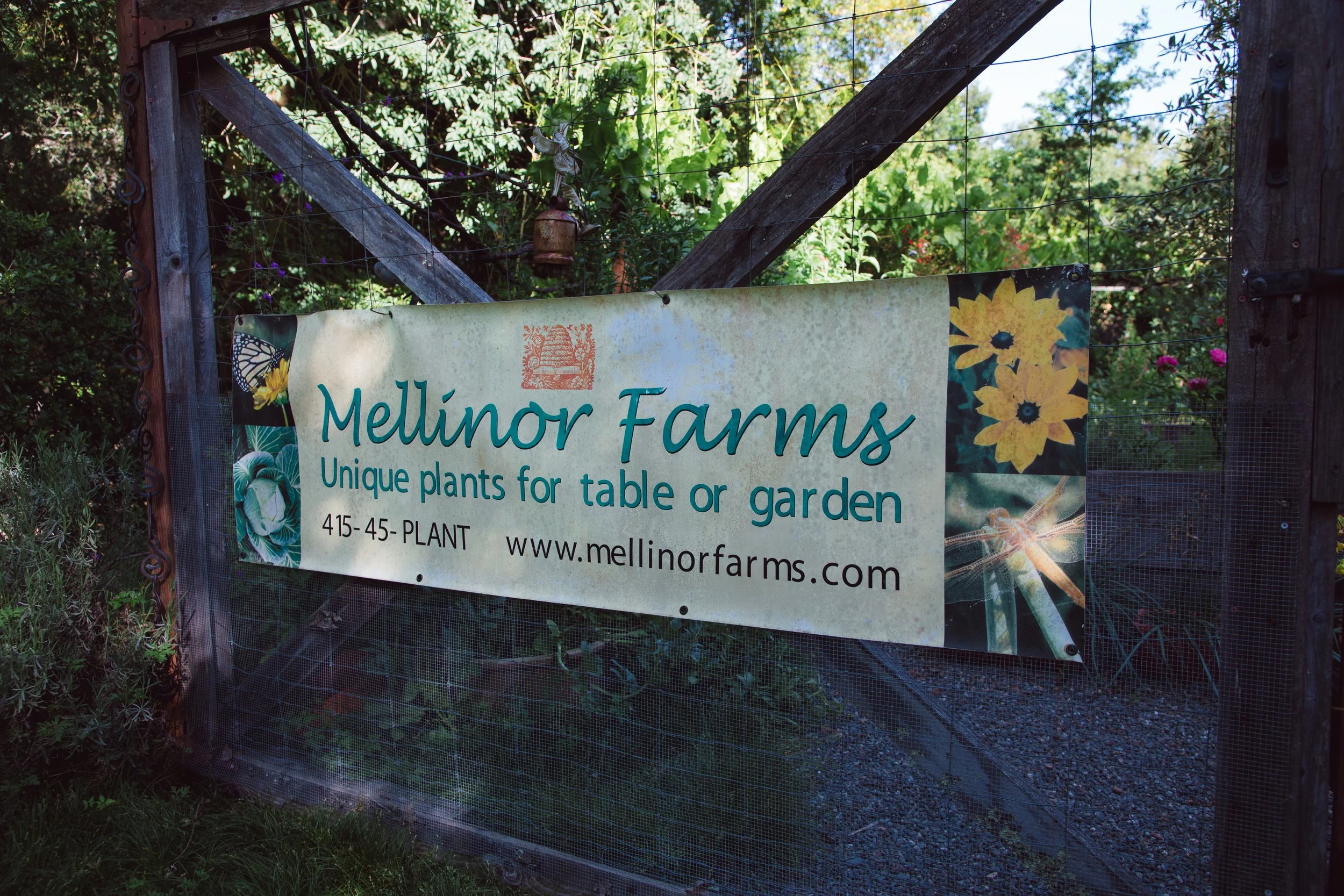 A farm sign reading "Mellingor Farms" with botanical illustrations of flowers and insects, mounted on a wooden gate surrounded by green foliage.