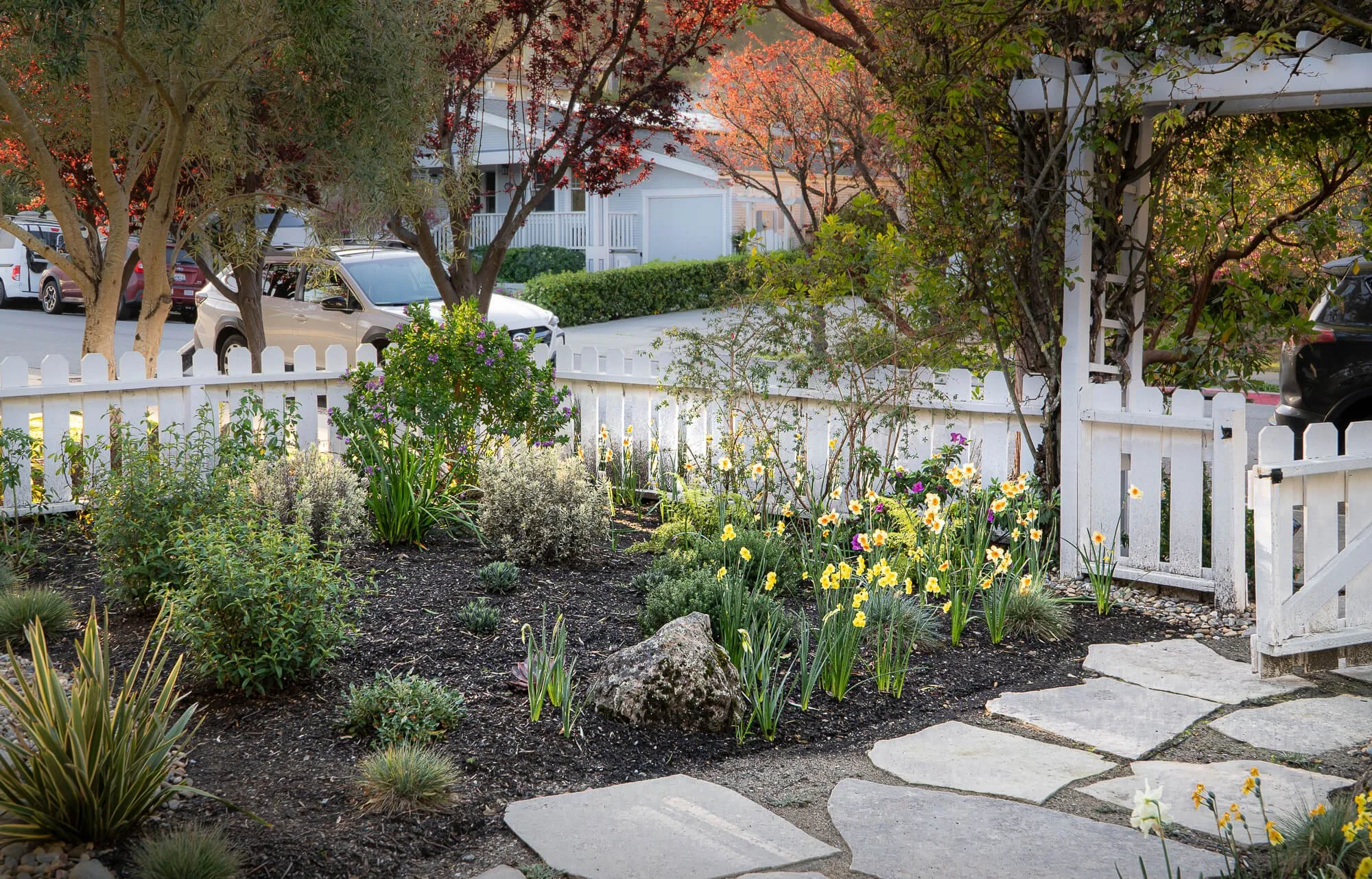 Front yard garden with flowering plants, green shrubs, and trees enclosed by a white picket fence, with a stone pathway leading to a garden gate.