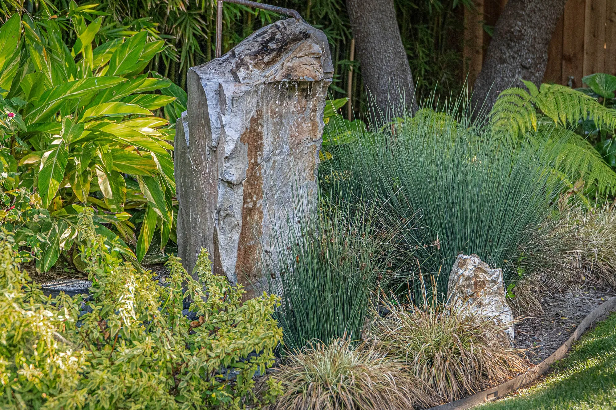 A garden bed with green foliage, including a large stone fountain, tall grasses, and leafy shrubs.