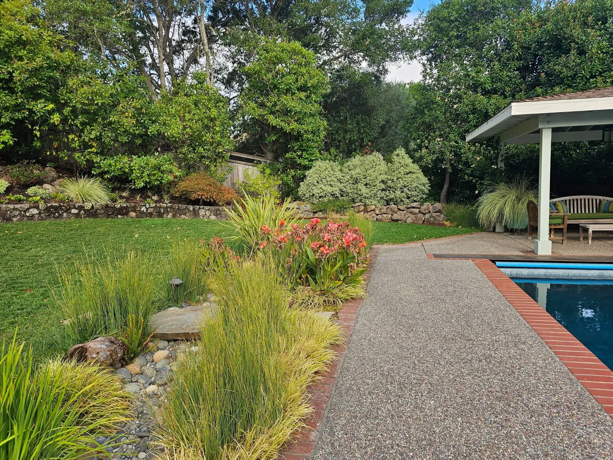 A backyard area with a garden, bushes, and trees on the left, a grassy lawn, and a patio with a swimming pool on the right. The patio has a seating area under a pavilion.