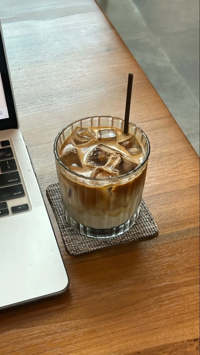 A glass of iced coffee on a woven coaster on a wooden table, next to a laptop.