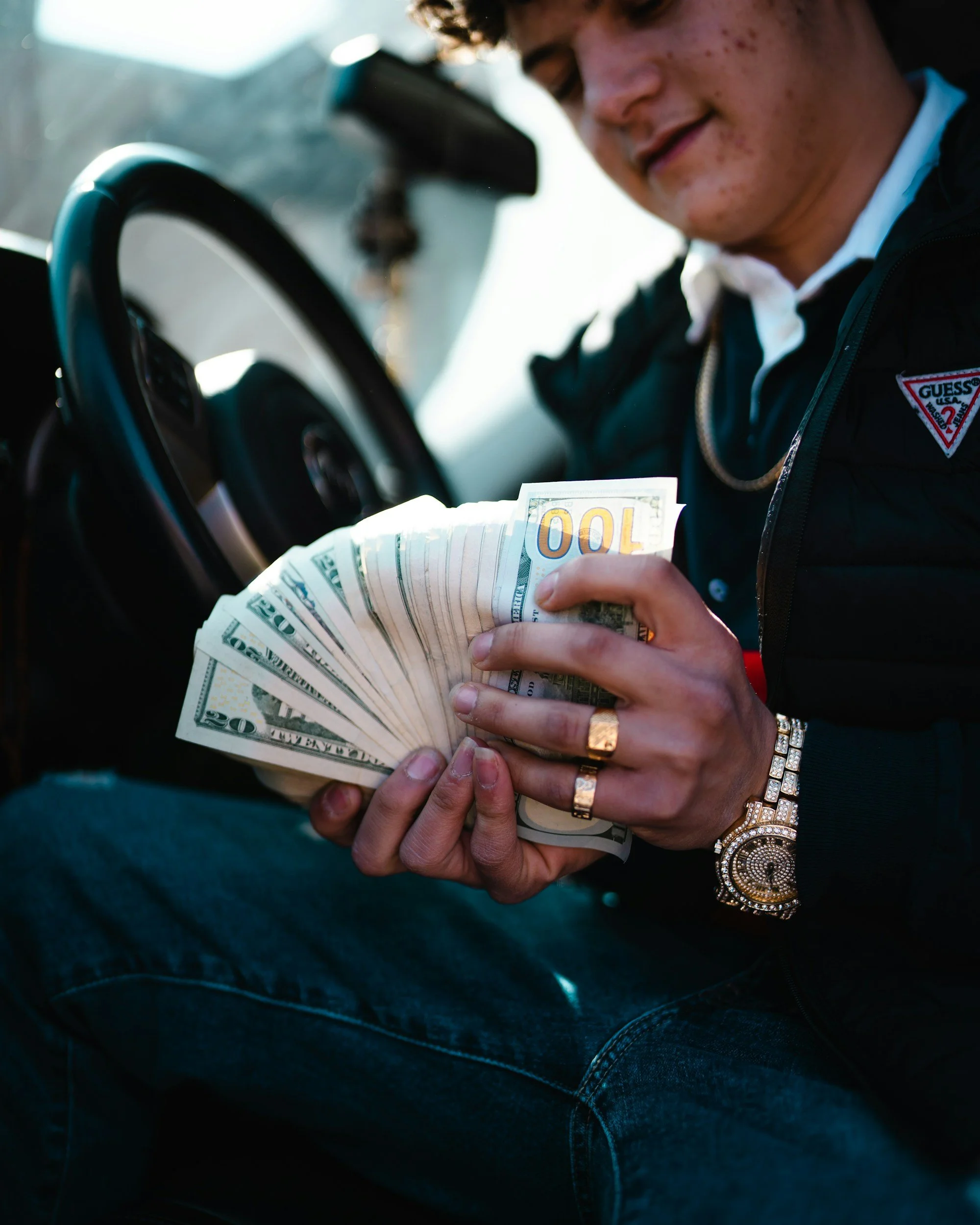 Person showing a fan of hundred-dollar U.S. bills inside a car with a steering wheel visible.