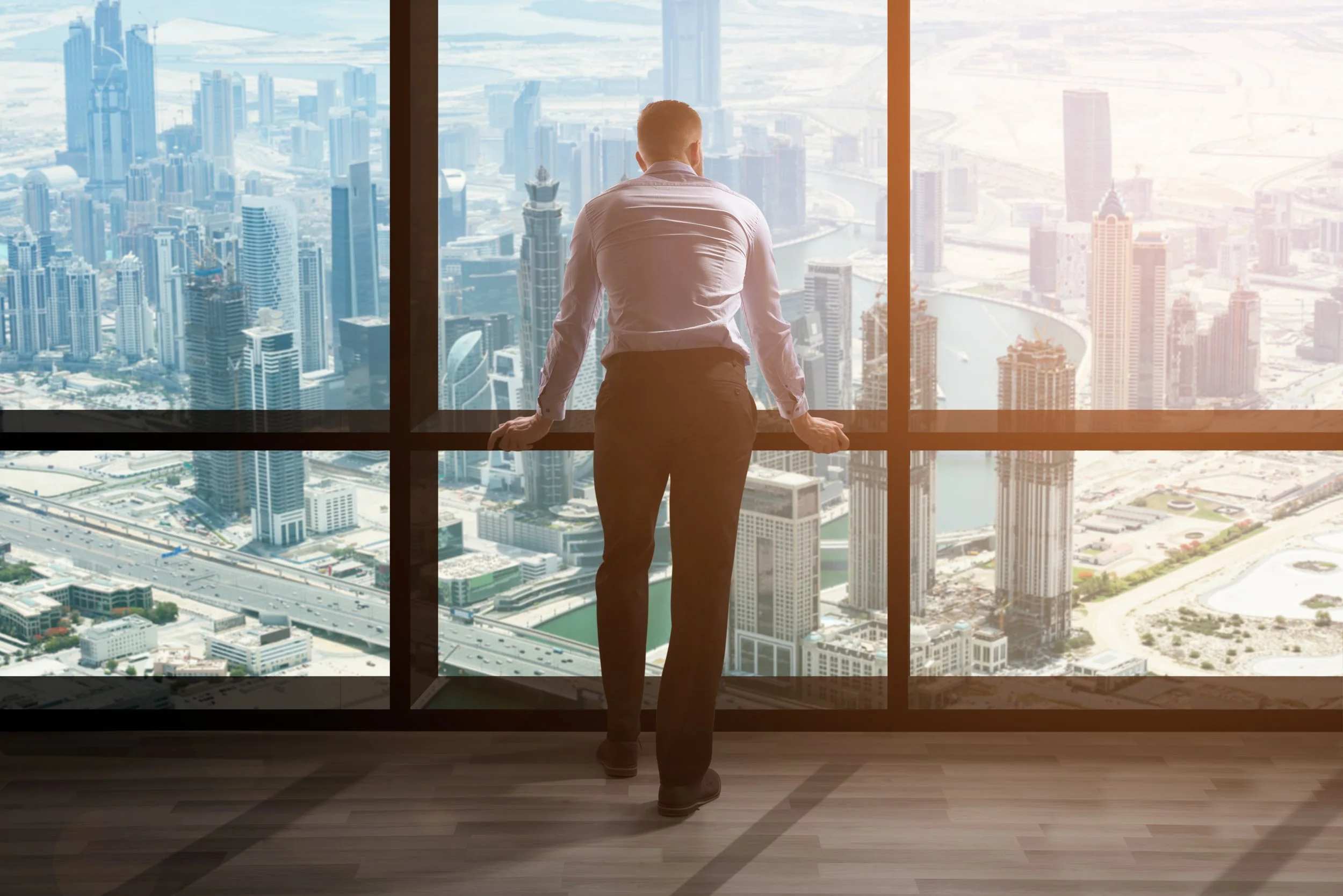 A man in a white shirt and dark pants standing inside a high-rise building, looking out of large window panels over a city skyline with numerous skyscrapers and a river.
