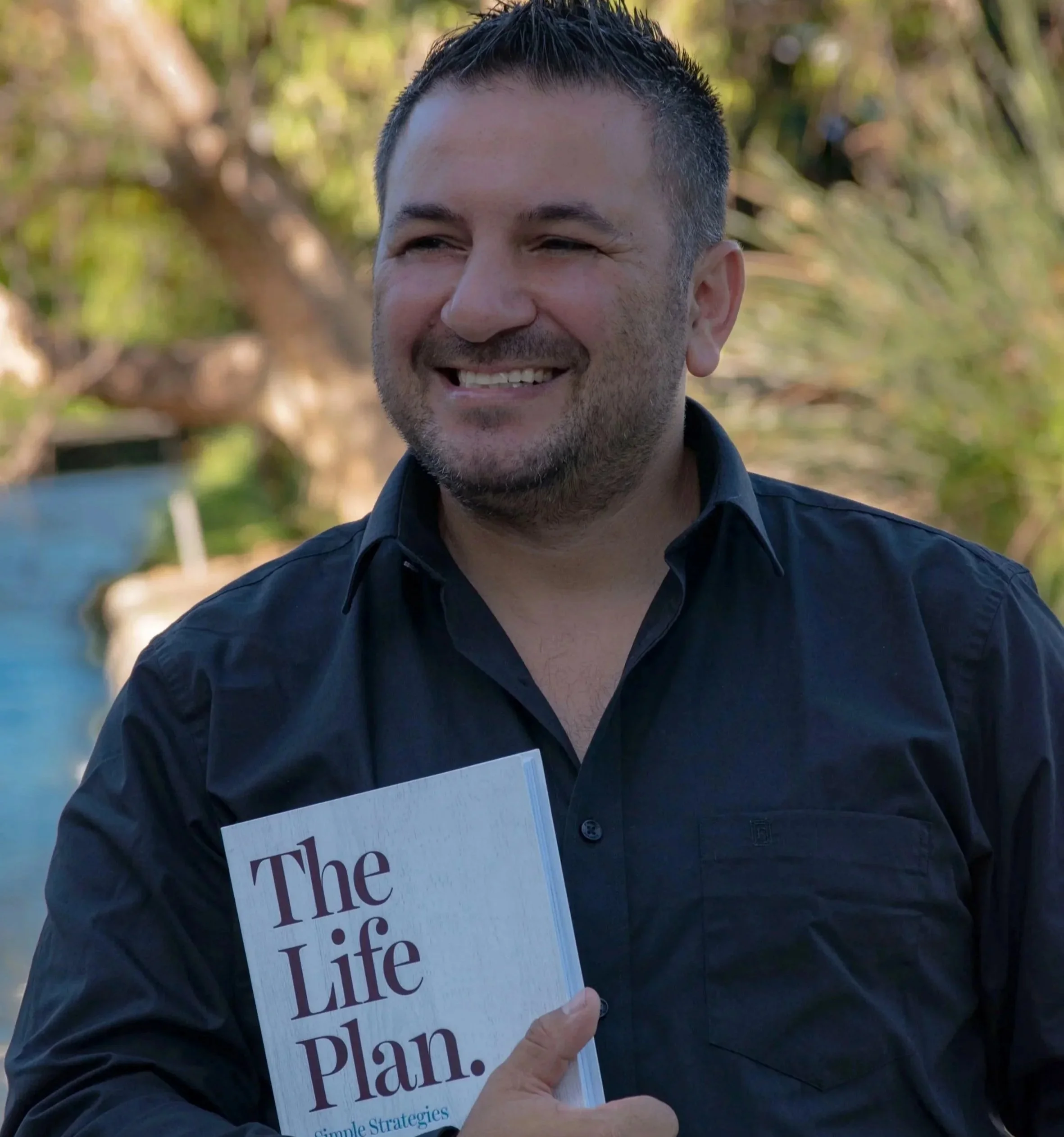 A smiling man in a black shirt holding a book titled "The Life Plan" outdoors with greenery in the background.