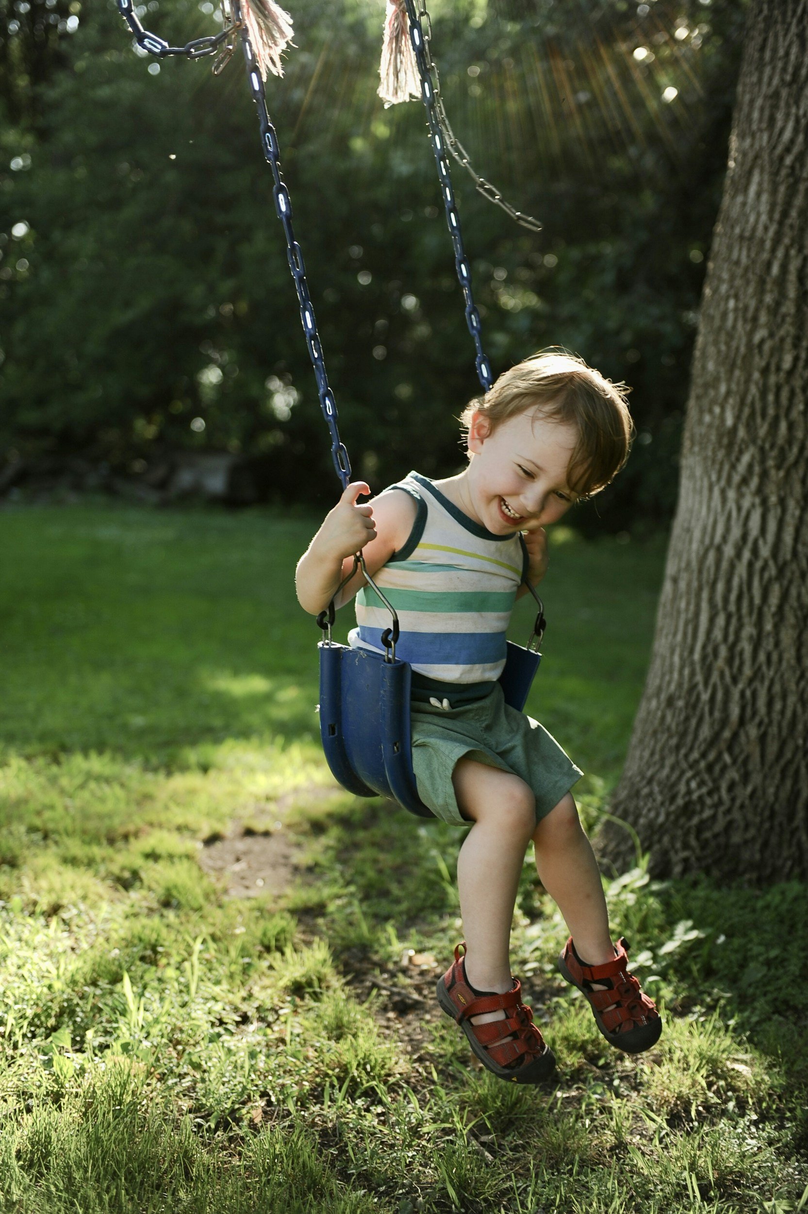 A young boy with brown hair, wearing a striped shirt and green shorts, is smiling and swinging on a blue swing set outdoors in a grassy yard with trees.