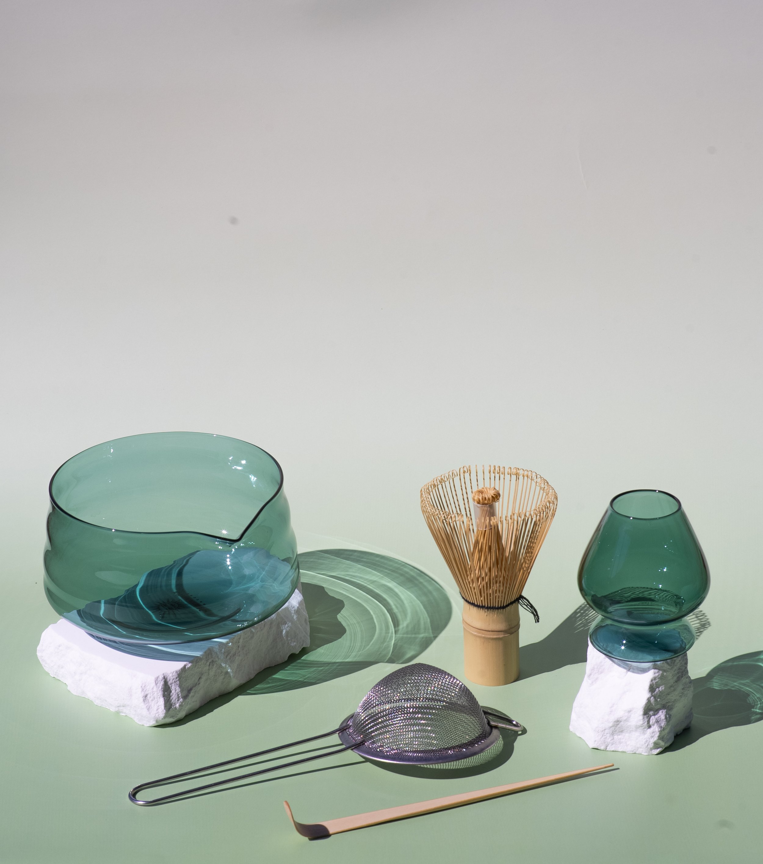 Collection of glass bowls and vases, a bamboo whisk, a metal tea strainer, and a bamboo spoon on a light green surface with a plain background.