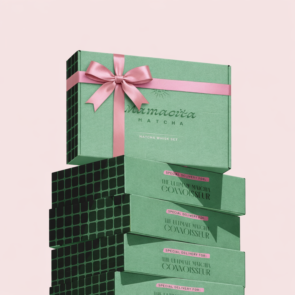 Stack of green matcha whisk sets in gift boxes, one with pink ribbon and bow, against a light pink background.