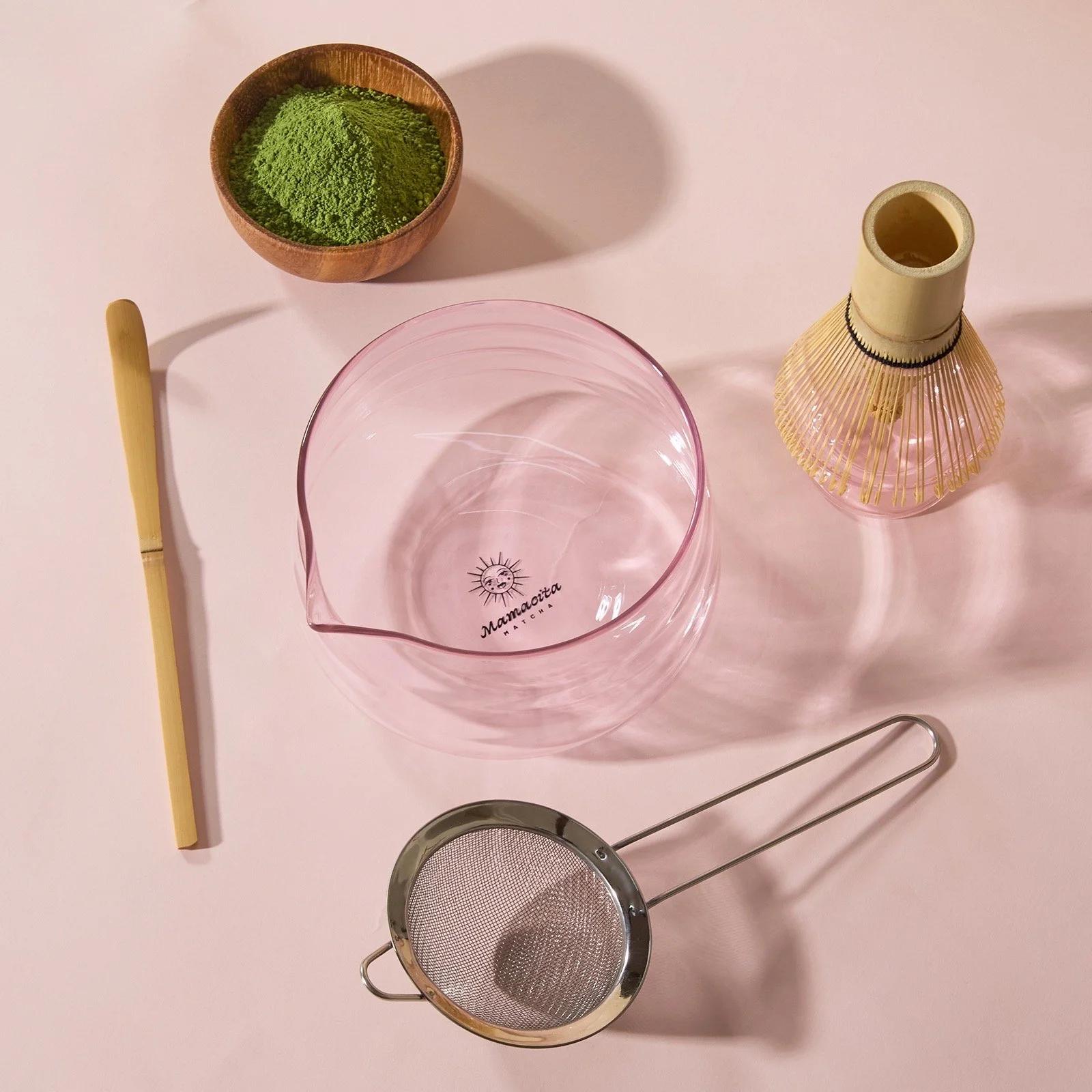 A decanter, a metal tea strainer, a small bamboo whisk, a pink glass pitcher with a sun and moon logo, a small bowl of green matcha powder, and a bamboo and rattan tea whisk holder on a pink surface.