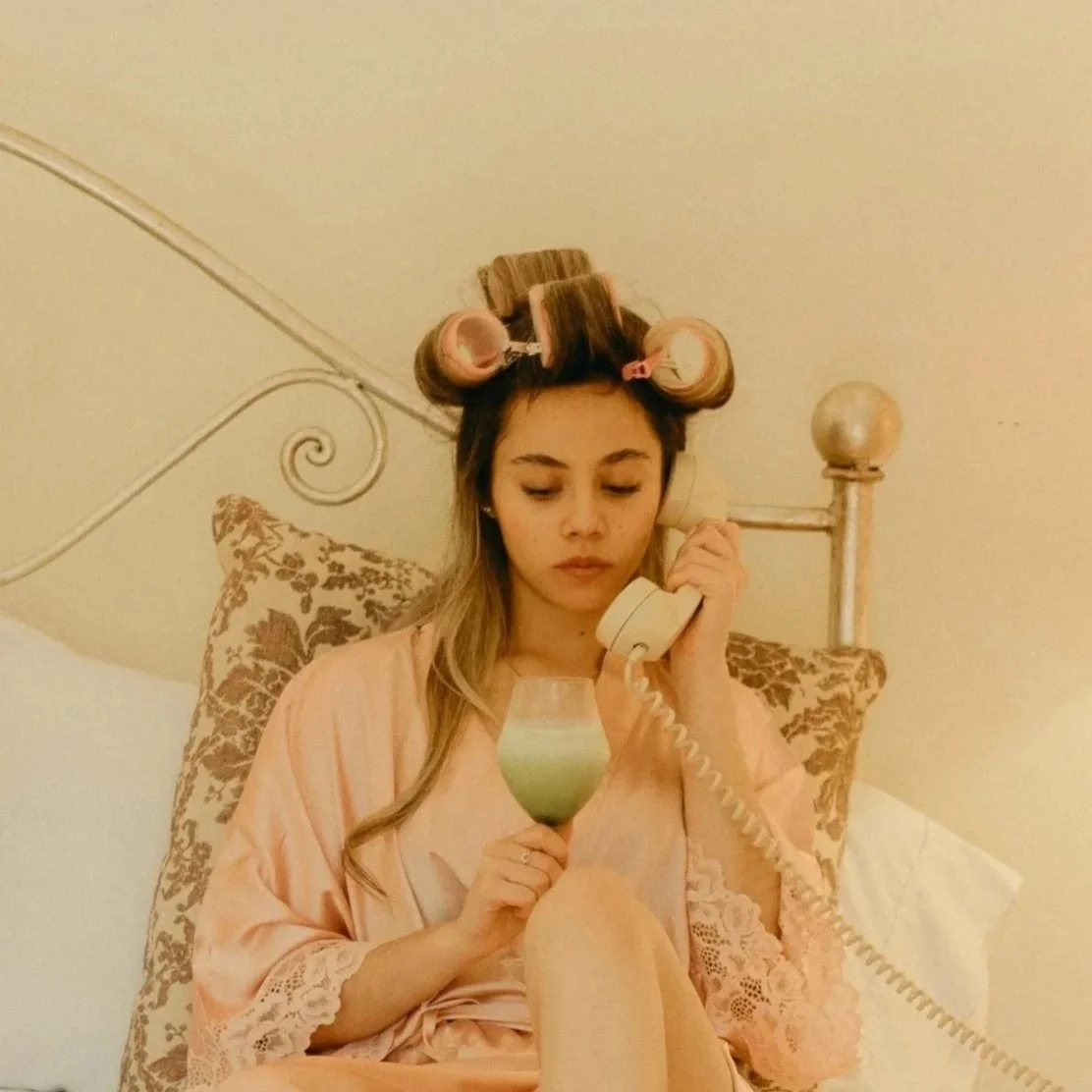 Young woman with hair rollers, sitting in bed, wearing a peach-colored satin robe, holding a vintage telephone receiver in one hand and a cocktail glass with a green drink in the other.