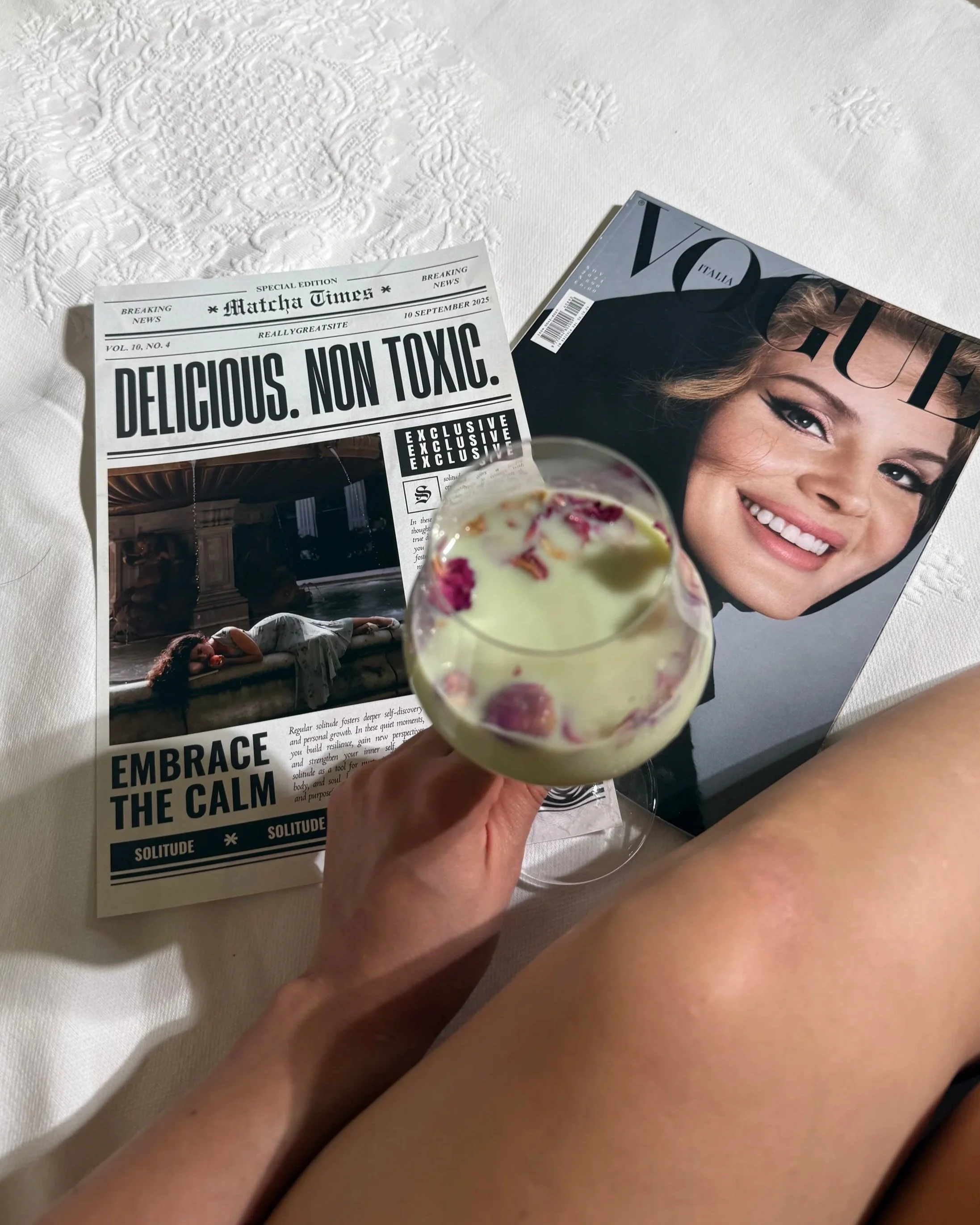 A hand holding a glass of light green cocktail with pink flower petals, on a white bedspread with embossed pattern. Nearby are magazines, one featuring a smiling woman on the cover and a newspaper with bold headlines.