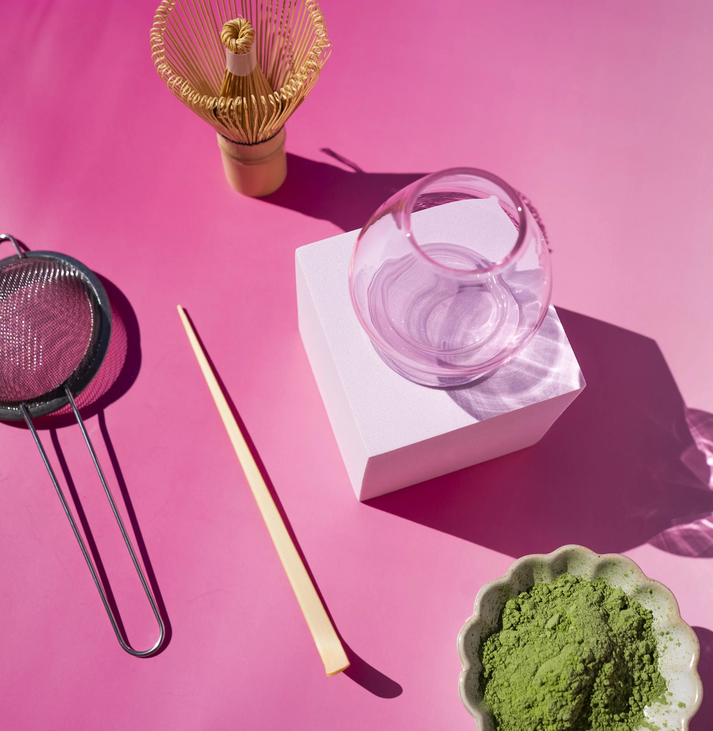 A pink background with various objects including a glass of water on a white block, a small bowl of green powder, a metal tea strainer, a wooden stick, and a gold-colored whisk-like object.